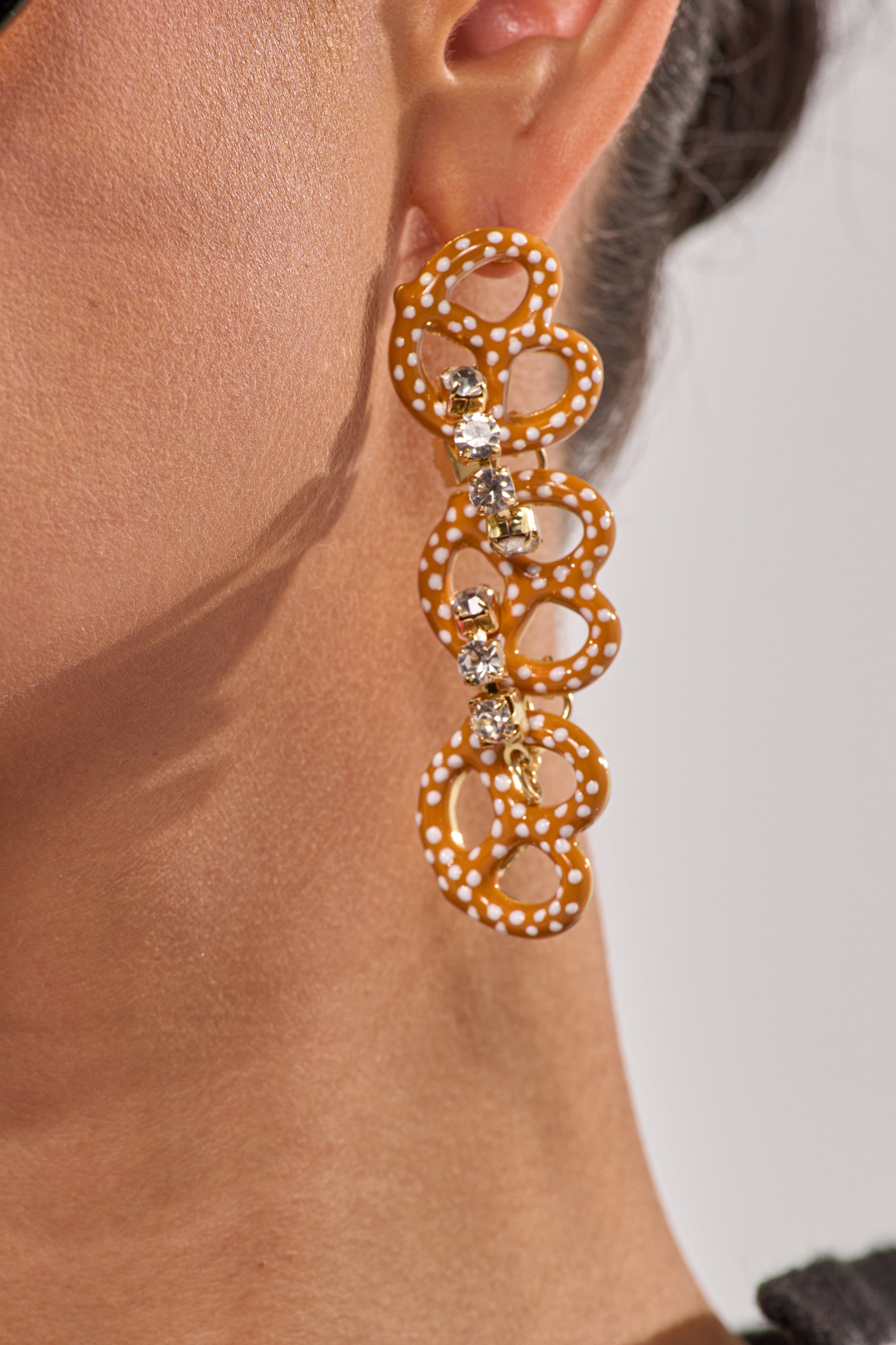 Close-up of a woman's ear wearing the NEVER SALTY EARRING—playful pretzel-shaped statement earrings with orange loops, white polka dots, and sparkling clear rhinestones, highlighted against her light brown skin and dark pulled-back hair. 5