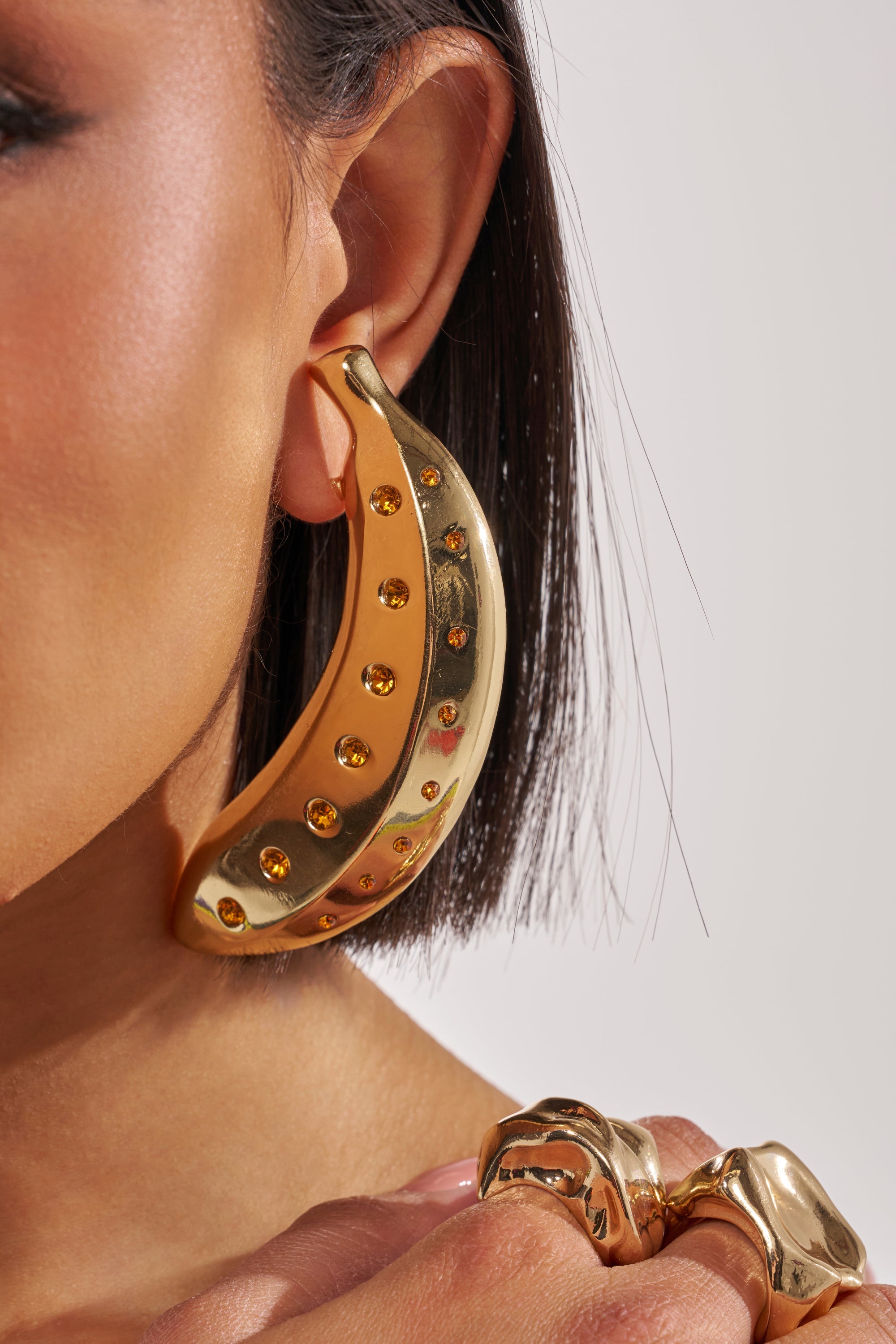 Close-up of a person wearing the GOING BANANAS EARRING—a bold, gold earring shaped like a banana with yellow gems. Their chunky gold ring and straight dark hair pop against the neutral background. 1