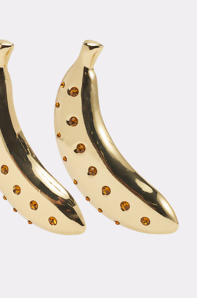 The GOING BANANAS EARRING features two gold banana-shaped earrings decorated with small amber gemstones, displayed side by side on a simple light background. 3