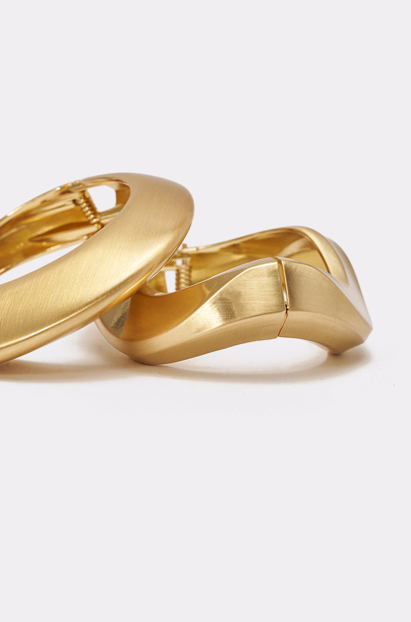 Close-up of the ULTIMATE STACK BRACELET SET: two gold-toned, sculptural bracelets with a smooth matte finish, one round and one with a wavy design, displayed on a light gray surface. Bold elegance inspired by chunky gold jewelry. 4