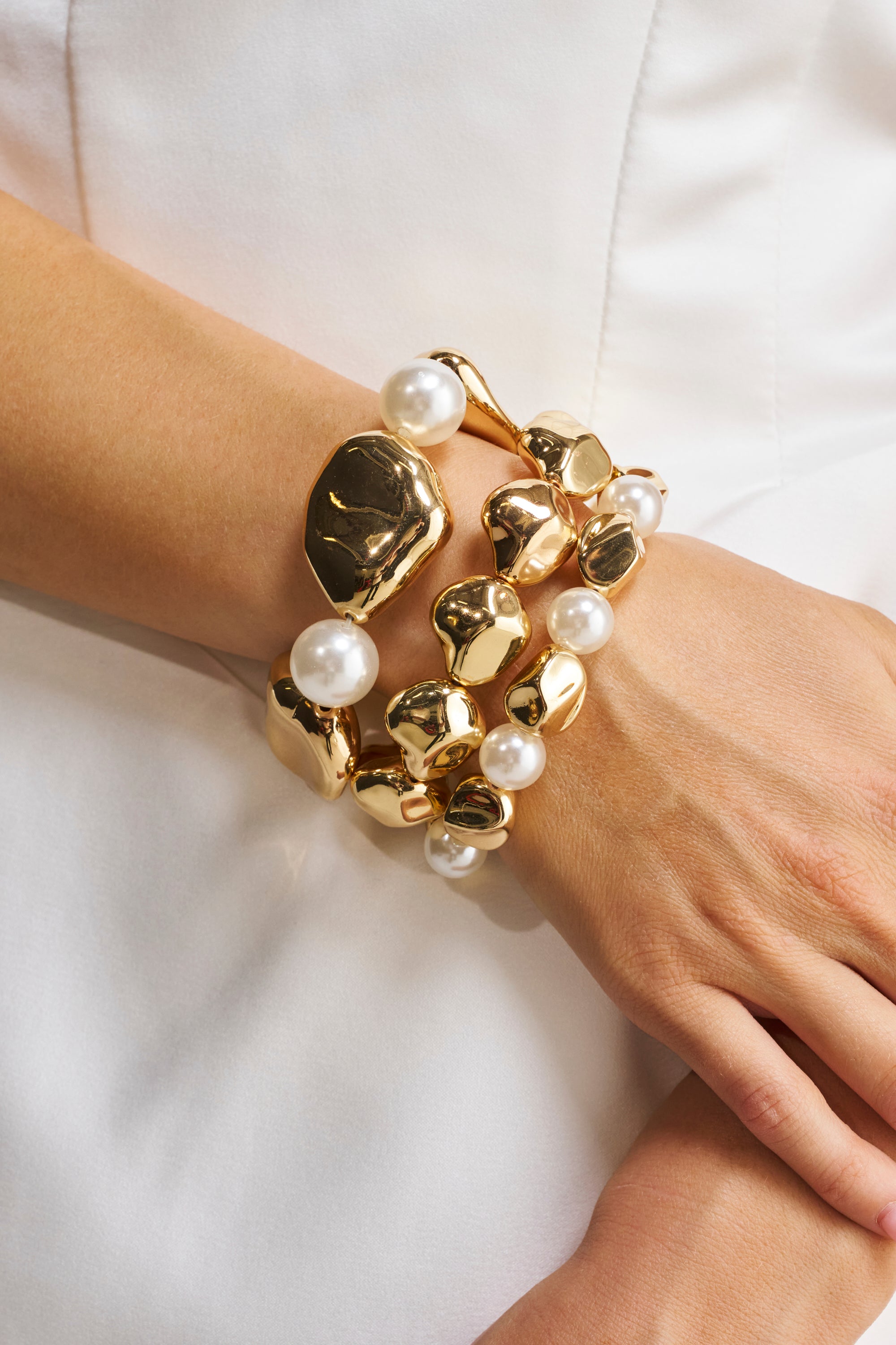 A close-up of a person’s wrist wearing the STACK IT UP BRACELET SET, featuring gold and faux pearl bracelets with large irregular gold pieces and white pearls, against a white fabric background.