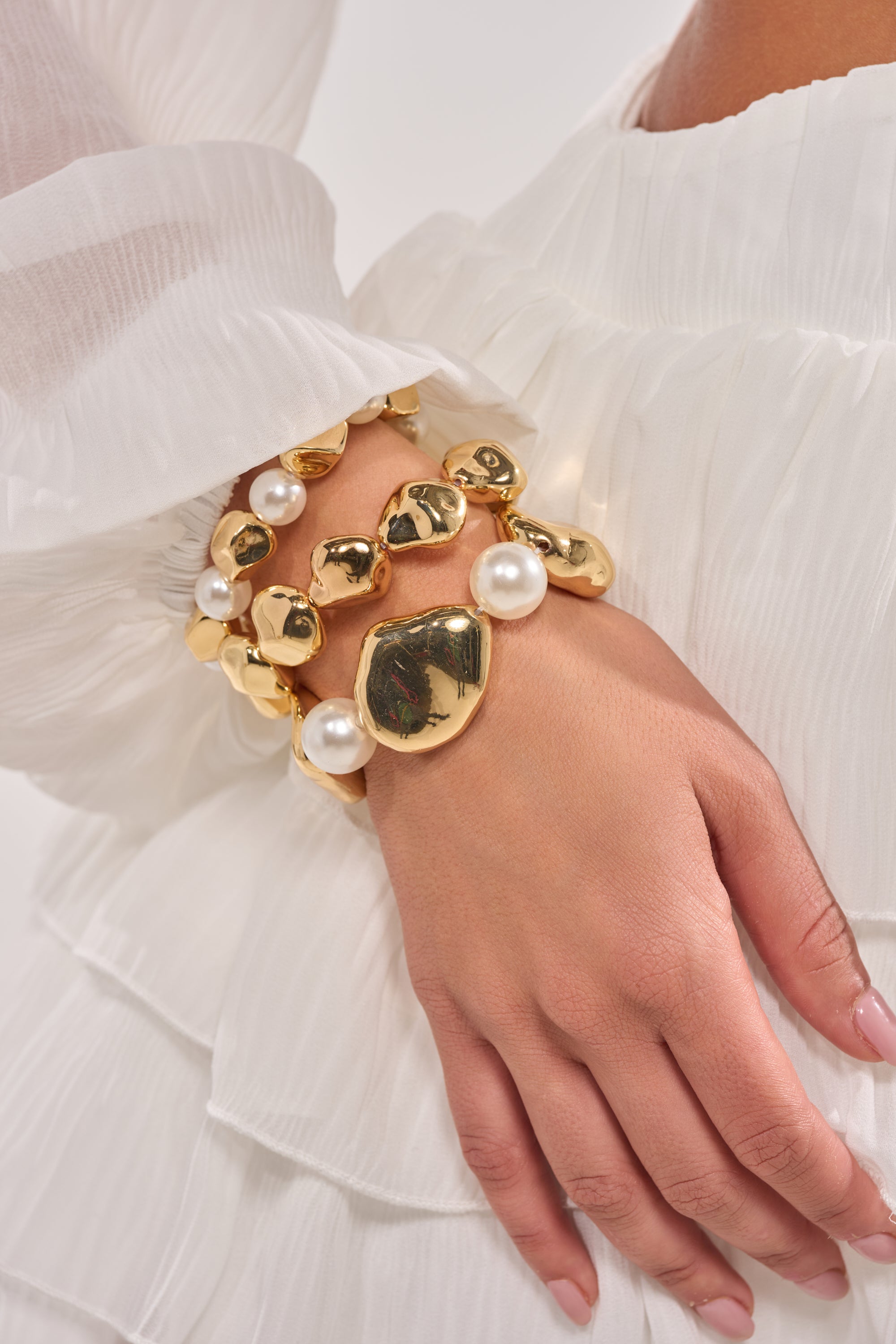 A close-up of a hand wearing the STACK IT UP BRACELET SET—three gold and faux pearl stretch bracelets—resting on a sheer, white, ruffled long-sleeve dress. The chunky statement set stands out against the delicate fabric. 1