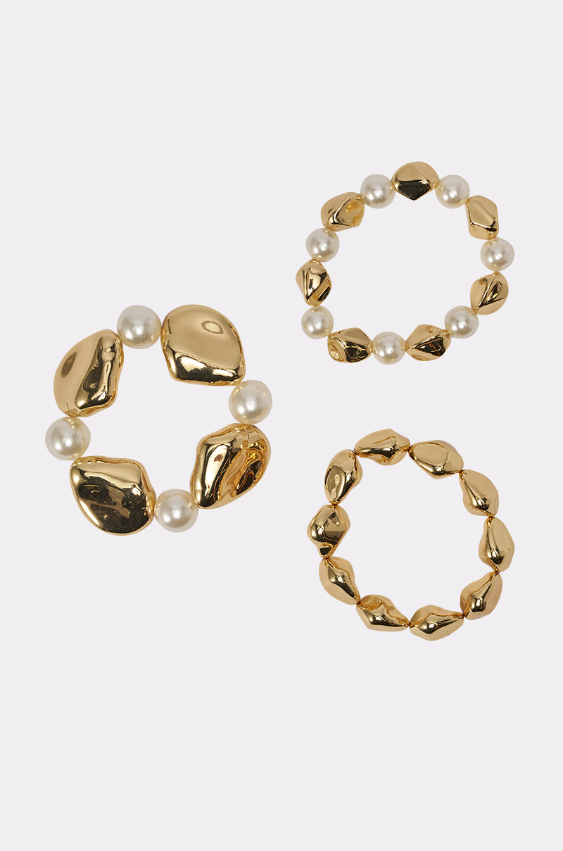 The STACK IT UP BRACELET SET features three gold-toned stretch bracelets: one with large irregular gold beads and faux pearls, and two with smaller gold beads—one alternating between gold beads and pearls—shown on a white background. 2