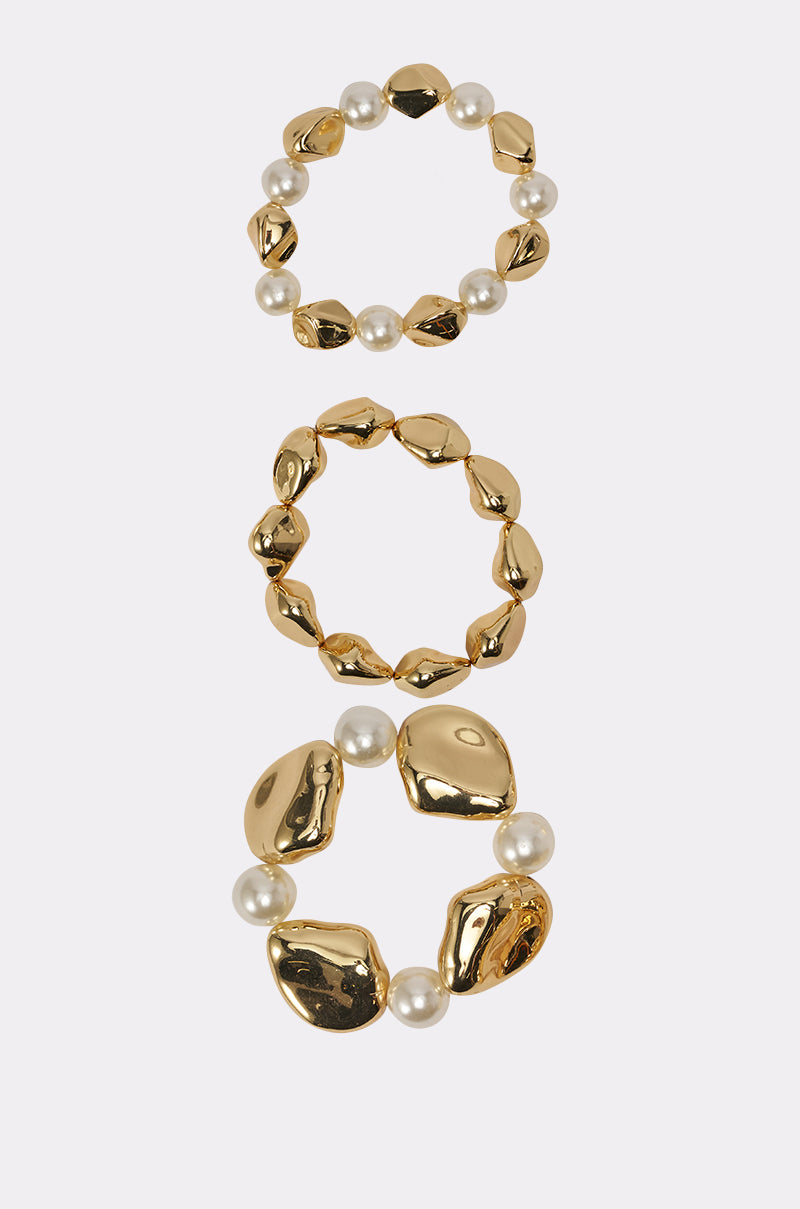 The STACK IT UP BRACELET SET features three chunky, gold and pearl stretch bracelets with irregular gold beads and round white pearls, arranged vertically on a white background for a bold statement look. 3