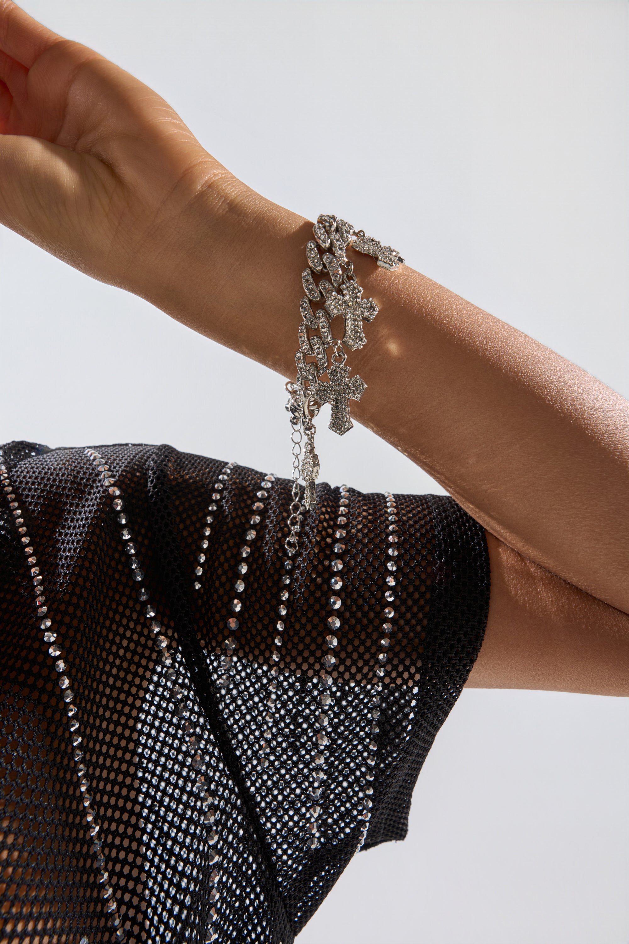 A close-up of a raised arm wearing a sparkling mesh top and the ICED OUT CHARM BRACELET, featuring chunky curb links with cross-shaped charms, set against a light background. 1