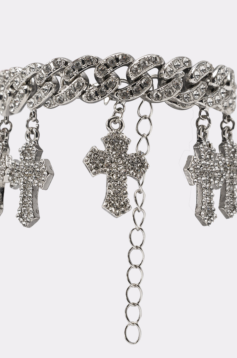 A close-up of the ICED OUT CHARM BRACELET, featuring a chunky curb-link chain embellished with sparkling rhinestones and multiple dangling cross pendants, shown against a plain white background. 3