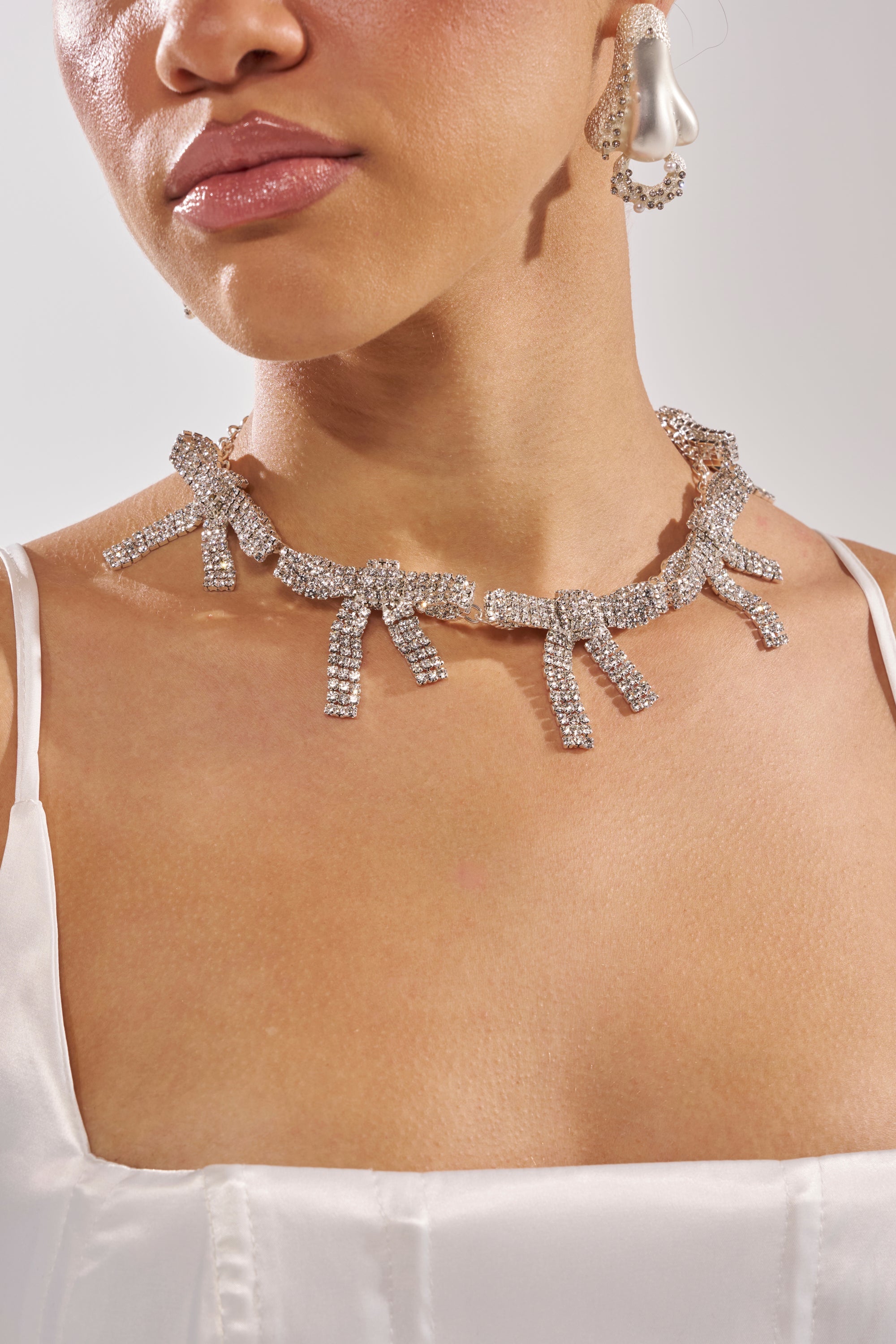 A woman wears a white satin top, IM THE GIFT NECKLACE—a statement bow necklace with rhinestones—and a large drop-shaped pearl earring, captured from the shoulders up against a plain background for an effortless coquette look. 0