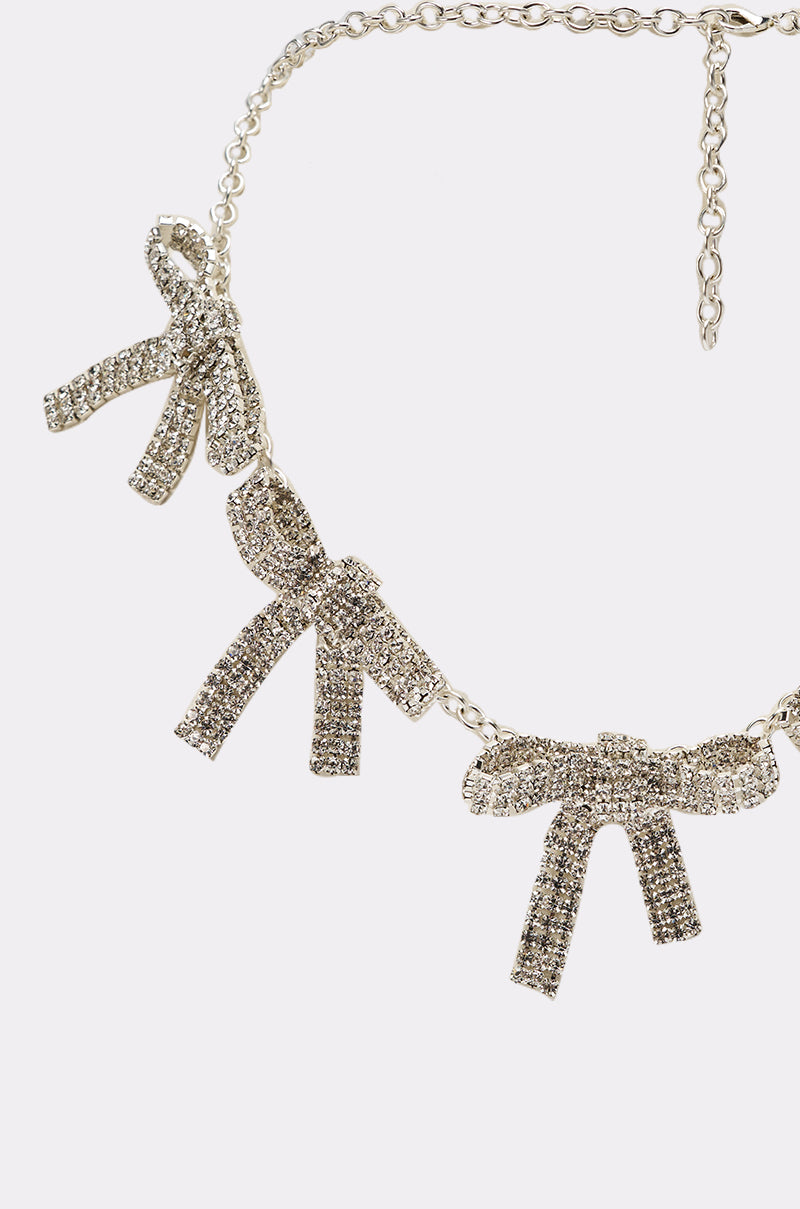 The IM THE GIFT NECKLACE is a silver statement piece with three bow-shaped pendants encrusted in sparkling rhinestones, shining brilliantly against a plain white background. 2