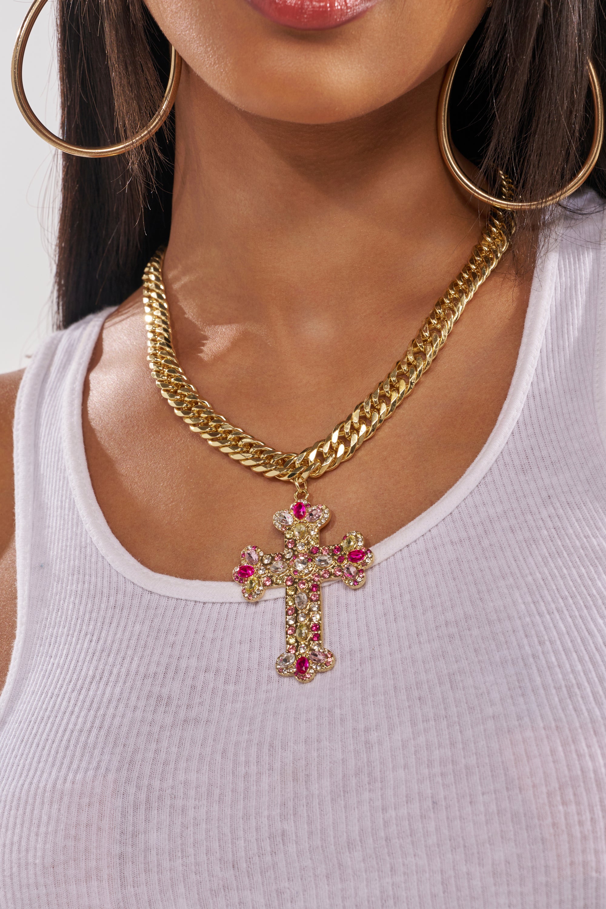 A woman wearing a white ribbed tank top, large gold hoop earrings, and the PERRY NECKLACE—a bold Cuban-link chain featuring a multicolor rhinestone cross pendant with pink and red stones. 1