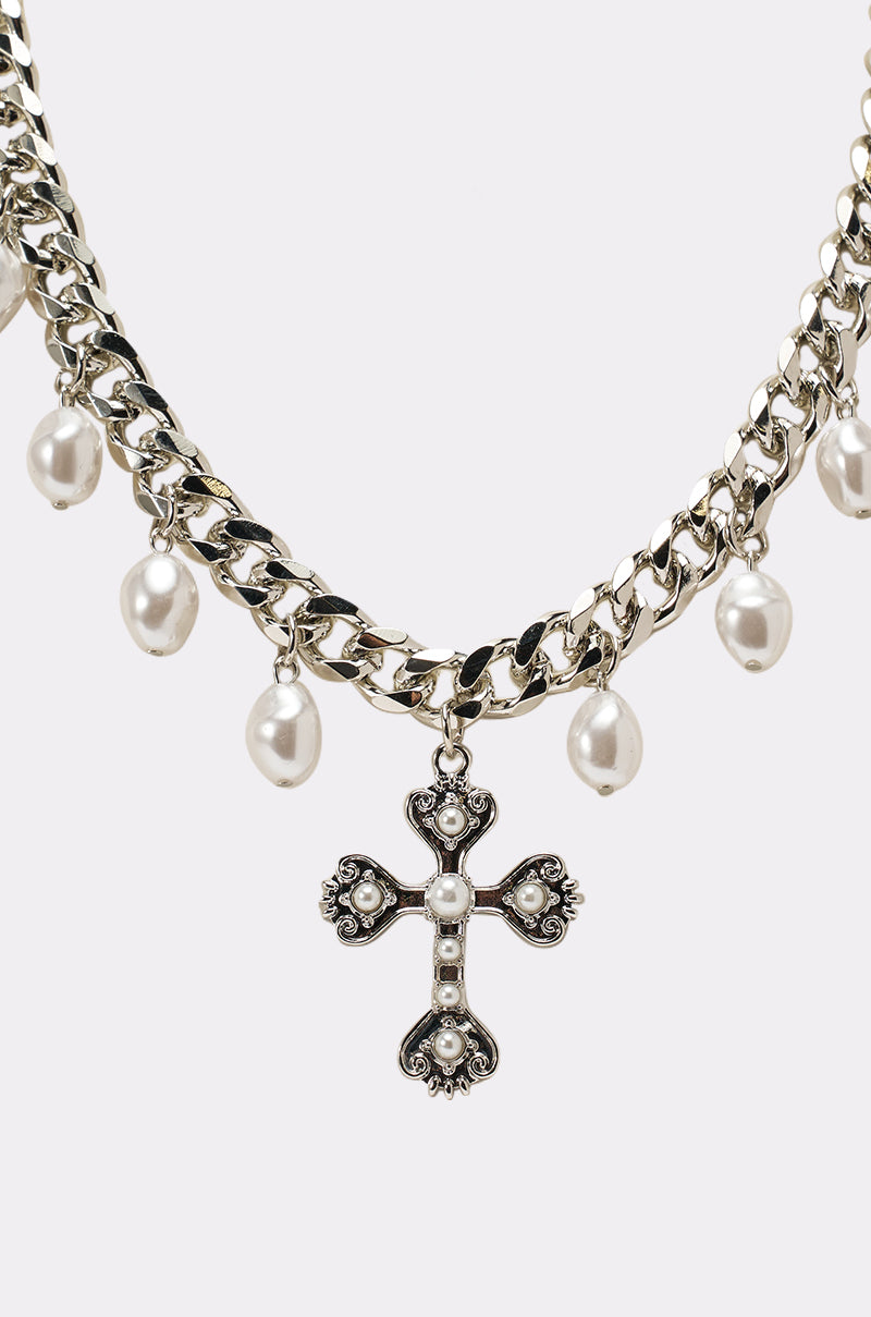 The TRINA NECKLACE is a silver Cuban-link chain featuring pearl-like beads and a decorative cross pendant with pearl-like accents, shown on a plain white background. 2