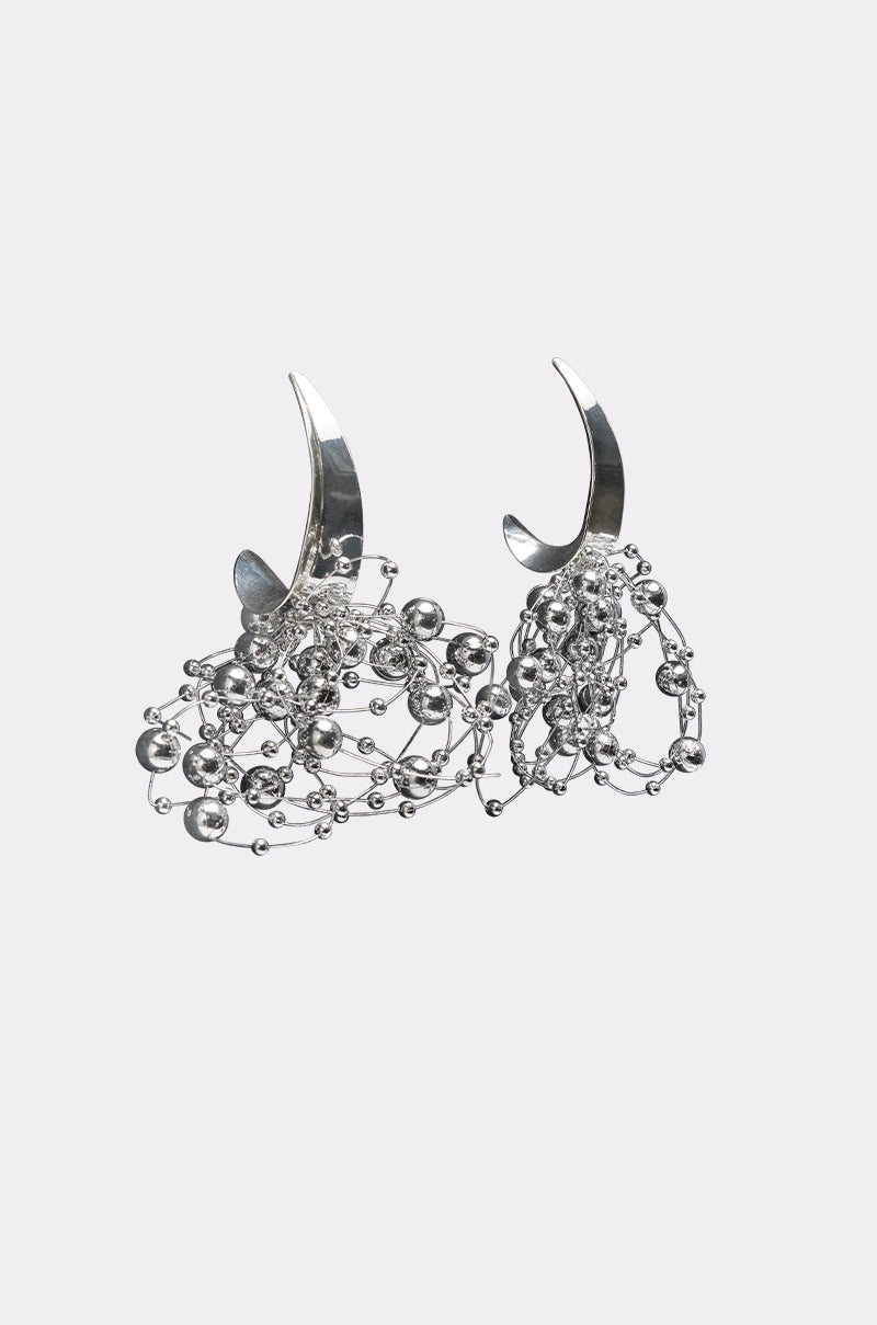 The BUBBLY EARRING is a pair of silver statement hoops with crescent-shaped hooks, featuring clusters of small metallic beads and chains, showcased against a plain light background. 0