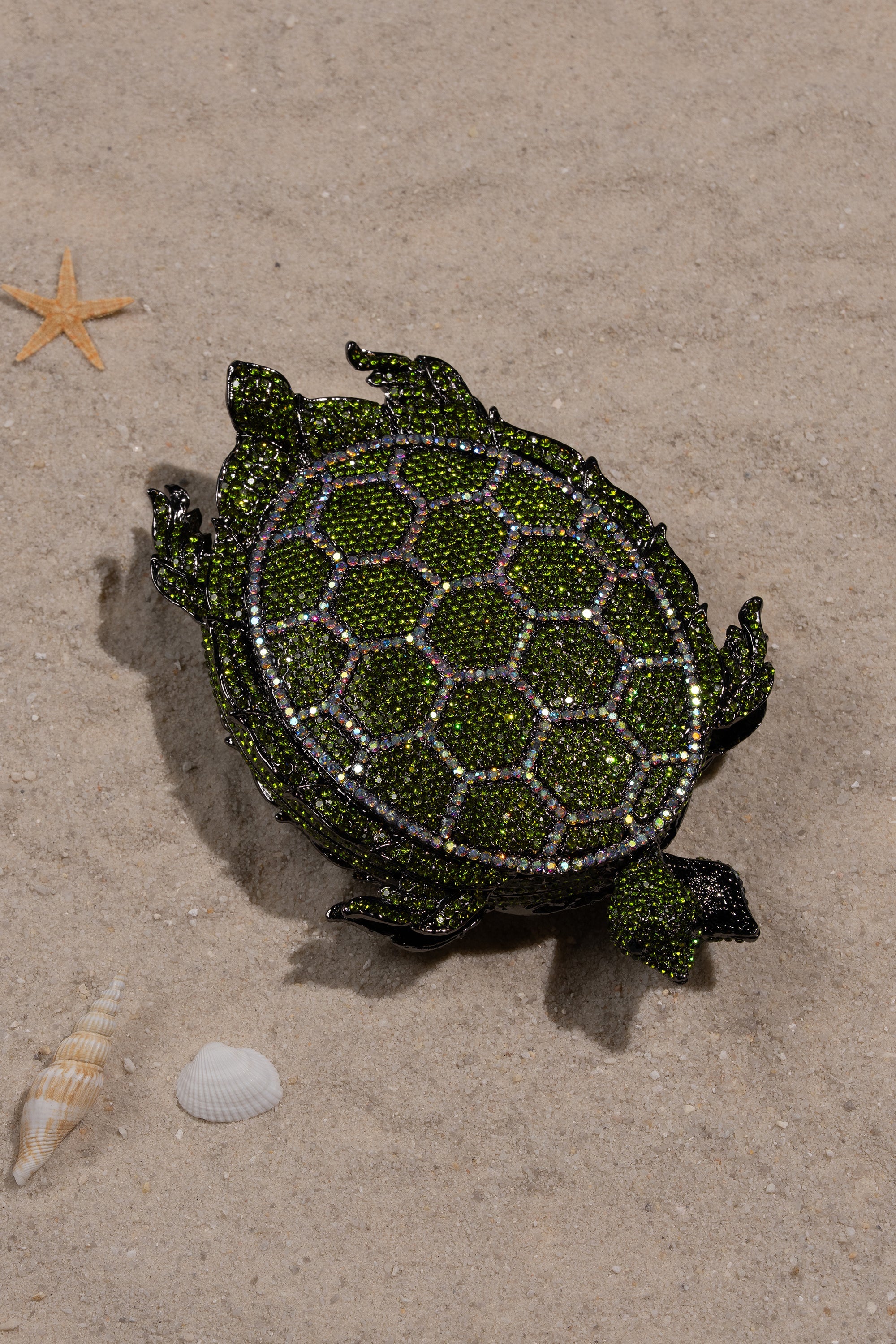 The BY THE SEA BLING PURSE, a jeweled turtle clutch with green and gold accents, sits on sandy ground by a seashell and starfish—an eye-catching statement handbag for any occasion. 1