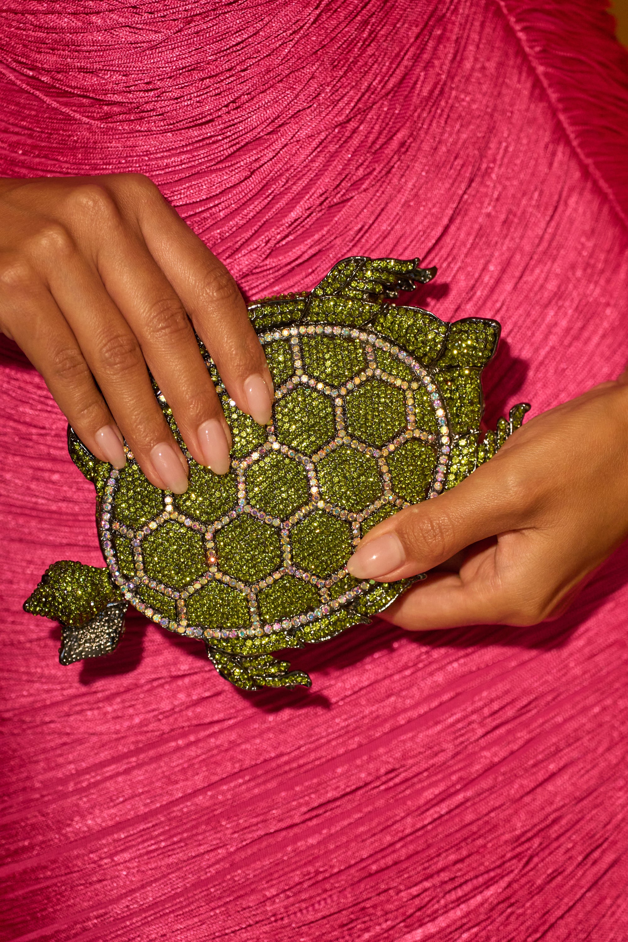 A person in a bright pink textured dress holds the BY THE SEA BLING PURSE—a statement evening bag shaped like a turtle, adorned with green and gold details that shimmer like rhinestones. 0