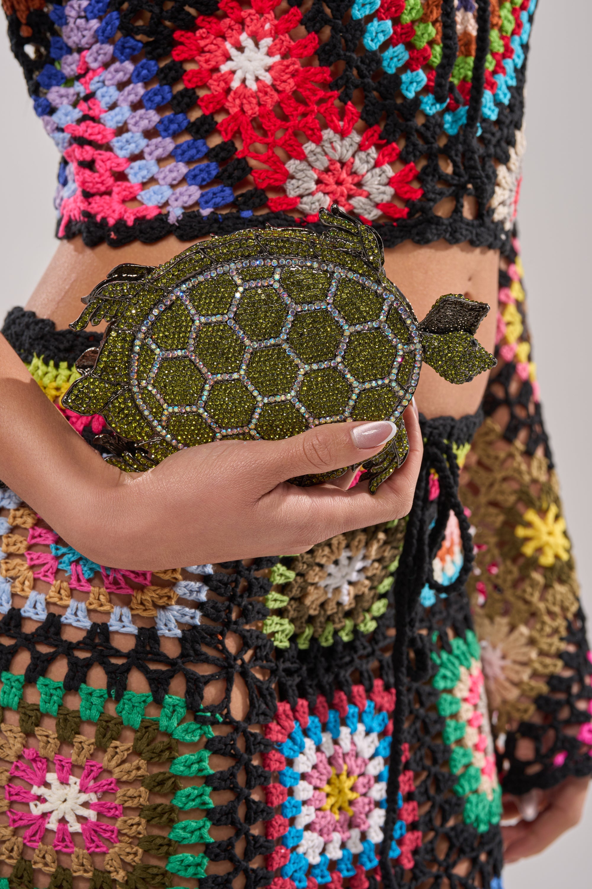 A person in vibrant crochet clothing holds the BY THE SEA BLING PURSE, a dazzling green rhinestone turtle-shaped evening clutch. The focus is on their hand grasping the purse and the intricate crochet patterns. 3