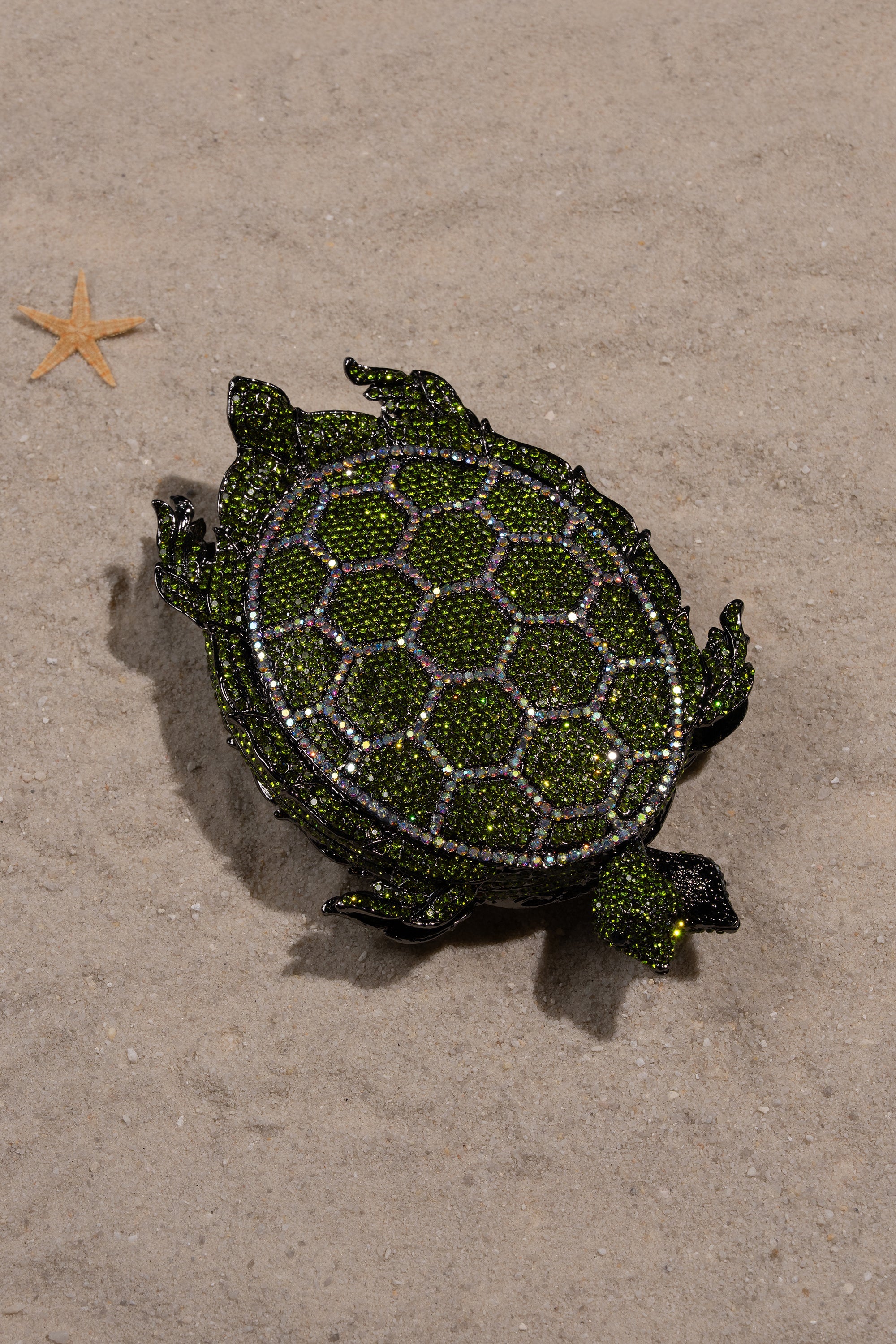 The BY THE SEA BLING PURSE, a bejeweled turtle clutch with green and gold rhinestones, rests on sand near a starfish—a stunning statement handbag. 6