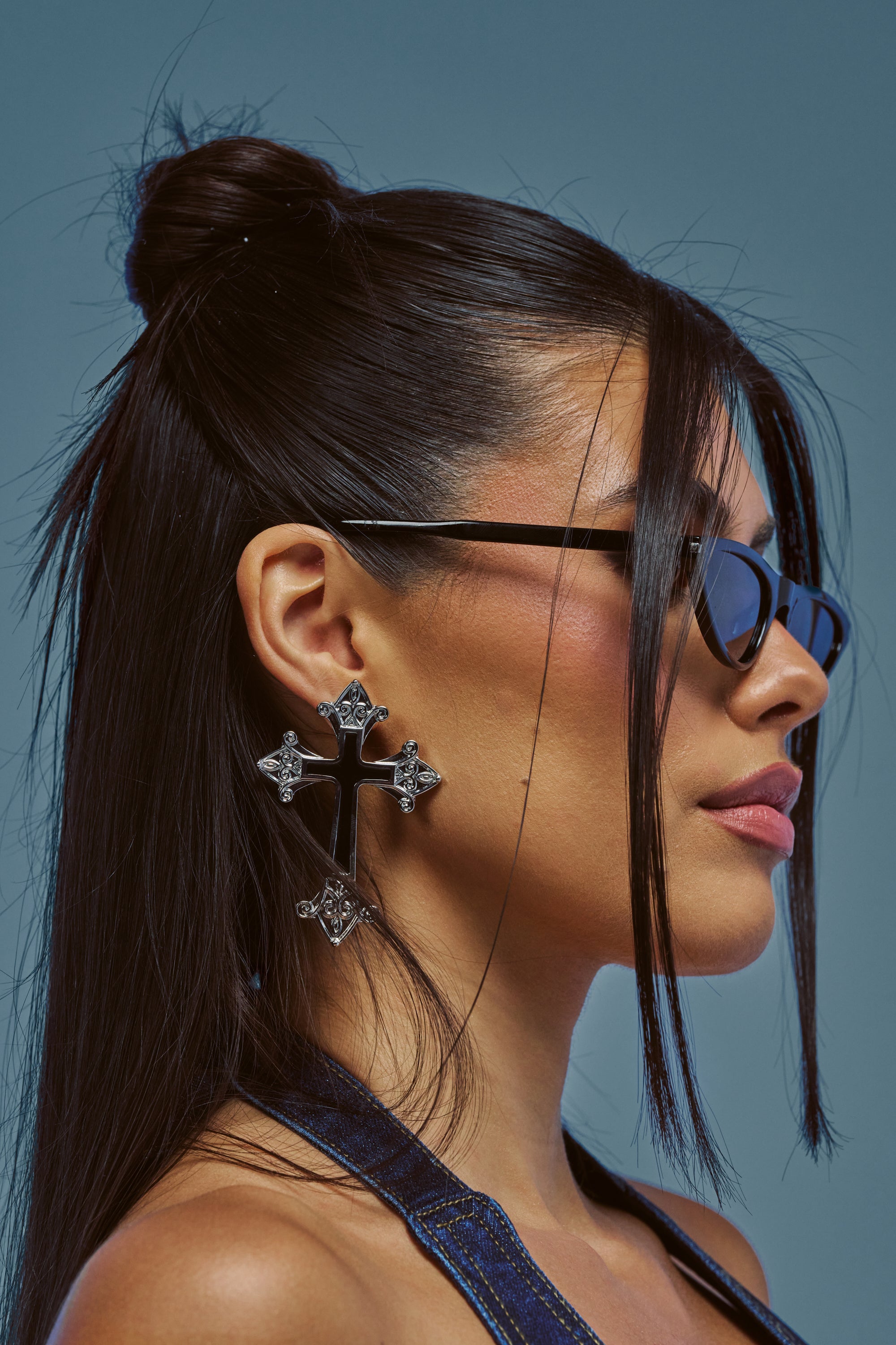 A woman with straight, dark hair styled half-up wears the ELITE ENERGY CROSS EARRINGS—large, ornate cross drop earrings—and black sunglasses as she looks to the side. She has glossy pink lipstick and a blue top.