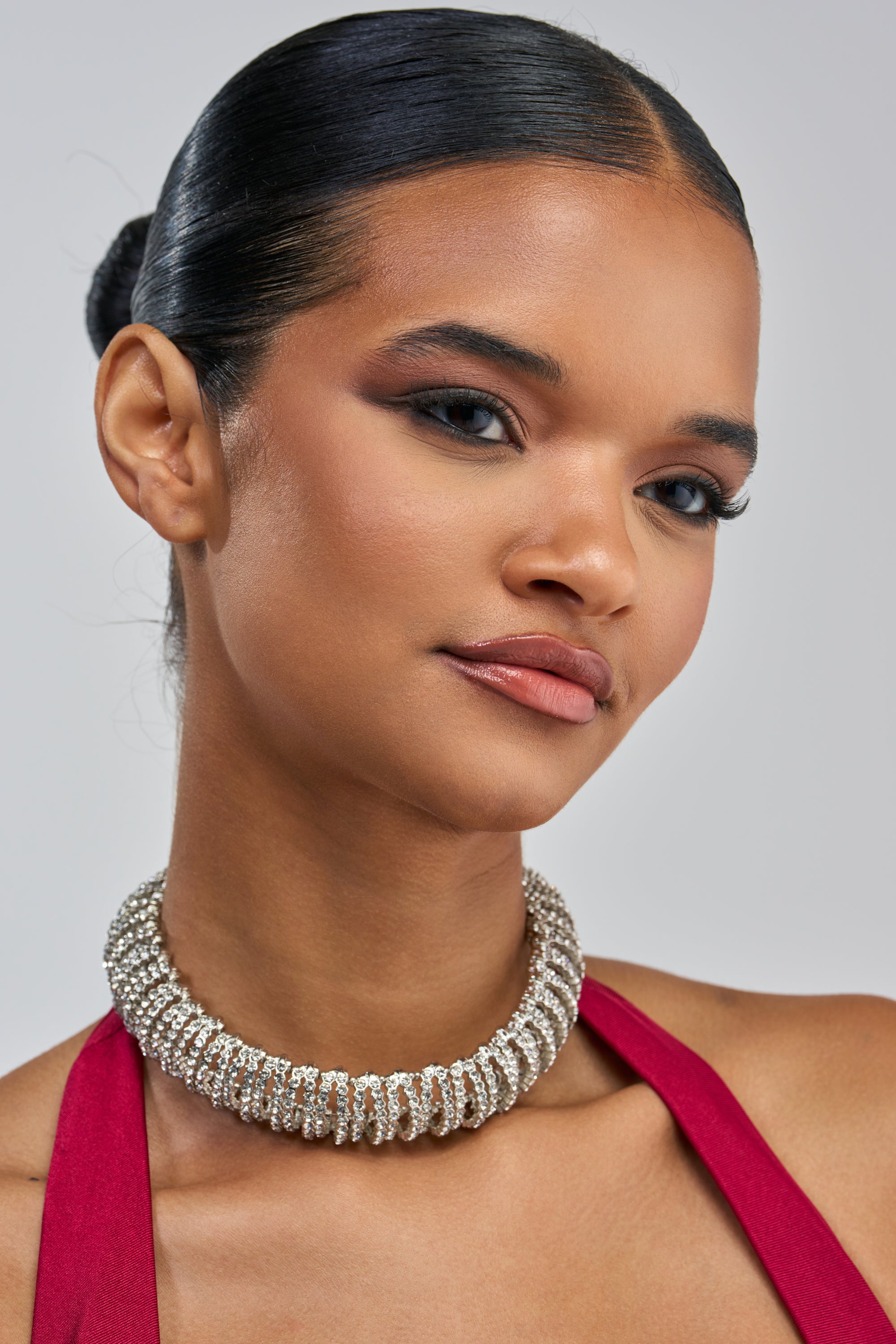 A woman with smooth dark hair styled in a bun wears the DIAMOND RUSH CHOKER, its silver rhinestones catching the light. She pairs it with a red halter top and soft makeup featuring bold eyeshadow and a neutral lip, gazing slightly to the side against a plain background.