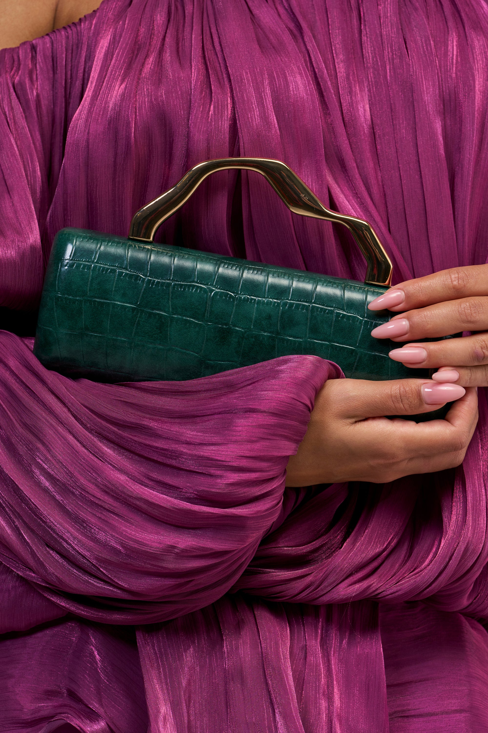 A person wearing a pleated magenta dress holds the ONE MORE TIME CLUTCH, a green crocodile clutch with a gold handle. Their nails are long and painted a light pink.