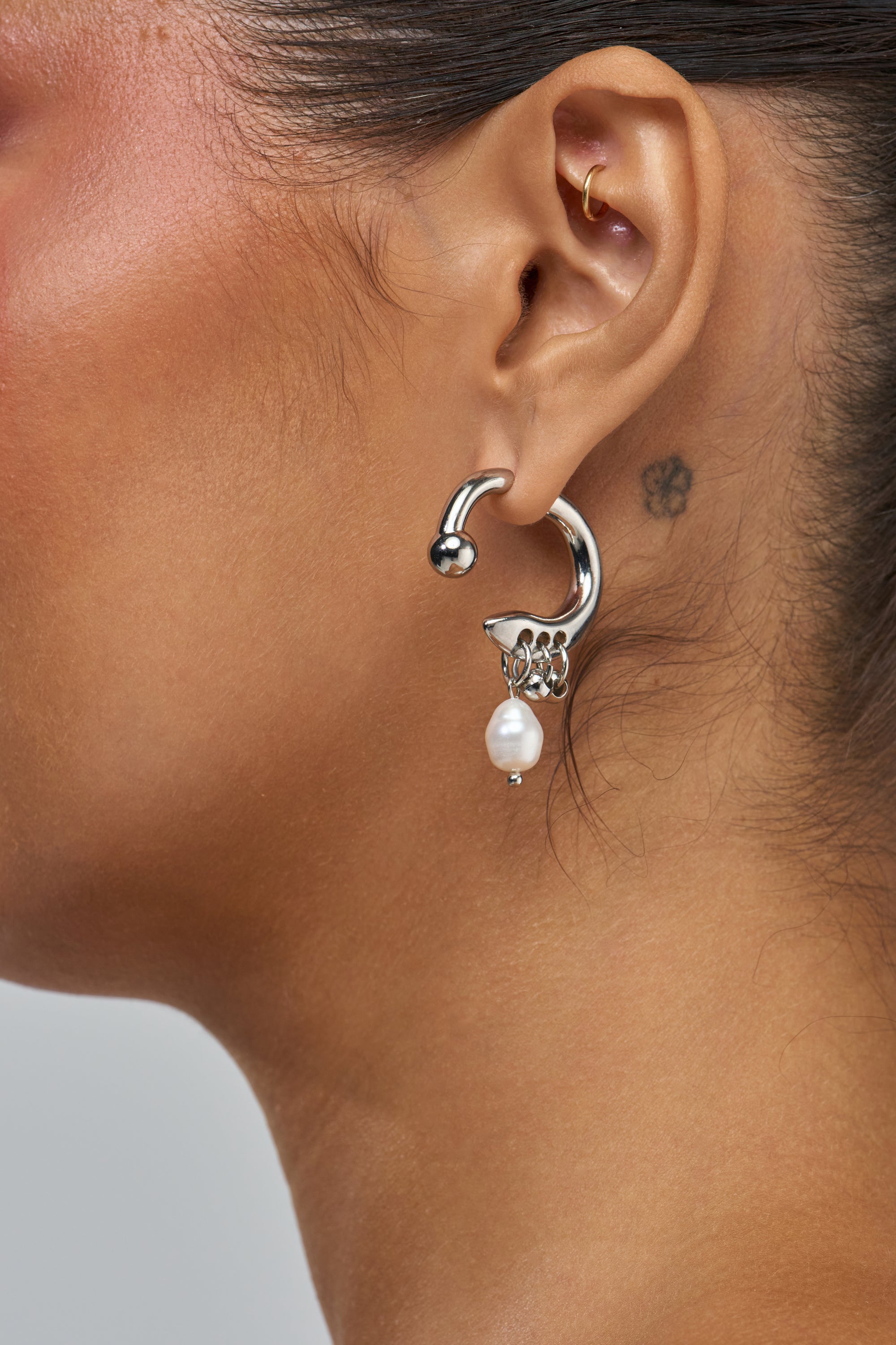 A close-up highlights a person’s ear adorned with gold cartilage hoops, the SHES SO MOD PEARL EARRINGS—featuring silver hoops with pearls and decorative details—and a small flower tattoo behind the ear. Their warm skin tone and pulled-back hair emphasize these elegant statement earrings.