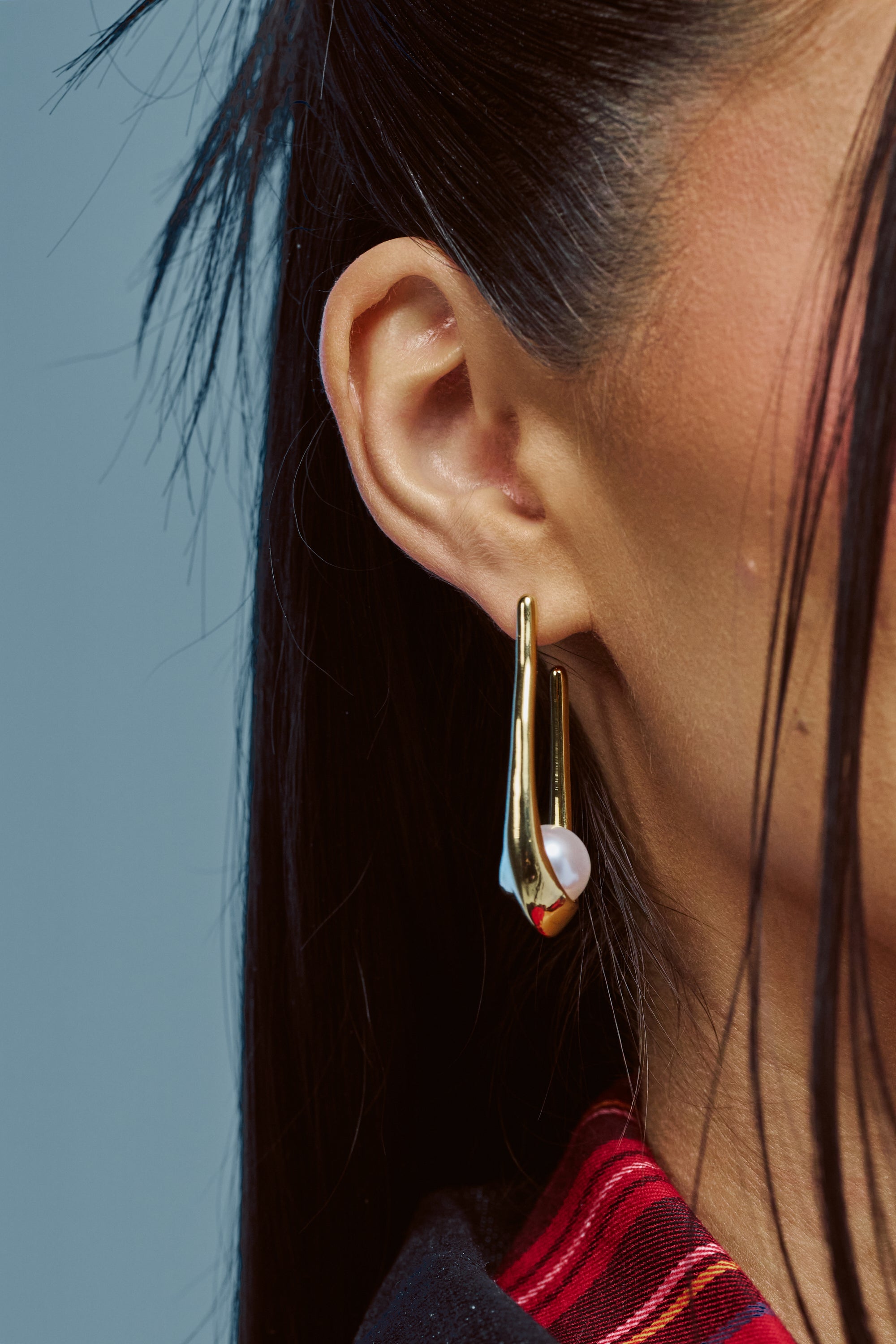 Close-up of a person’s ear with long, straight dark hair, wearing the HOLD MY PEARLS DANGLE EARRING IN GOLD—an abstract-shaped gold earring elegantly accented with pearls for a perfect bridal accessory. The collar of a red and black plaid shirt is partially visible.