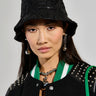 A woman models the RUNWAY BUCKET HAT, styled with a black jacket embellished with silver studs and white lettering. Her long dark hair pairs beautifully with pearl earrings and a necklace, while the jacket is highlighted by green and white accents against a neutral backdrop.