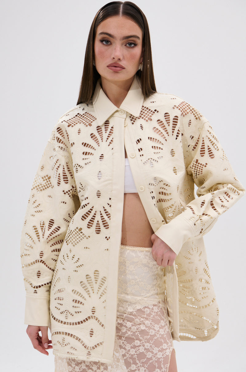 A woman wears the YOU FEELIN ME NOW CUTOUT PU BUTTON UP SHIRT, a cream long-sleeve top with cutout details, layered over a white crop top and lace skirt, standing against a plain white background.