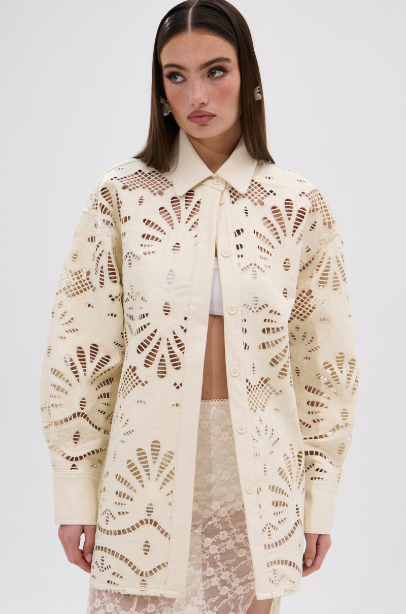 A woman wears the YOU FEELIN ME NOW CUTOUT PU BUTTON UP SHIRT—a long, cream-colored lace top with cutout detailing and a floral-inspired pattern—layered over a white bralette and lace skirt, against a plain white background.