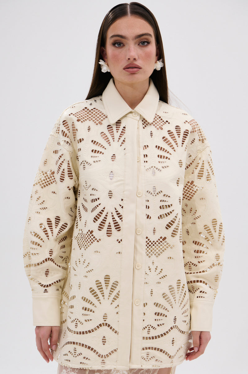 A woman with long brown hair wears the YOU FEELIN ME NOW CUTOUT PU BUTTON UP SHIRT, a cream-colored, long-sleeve blouse with floral-inspired cutout details. She stands against a plain light background and wears large floral earrings.