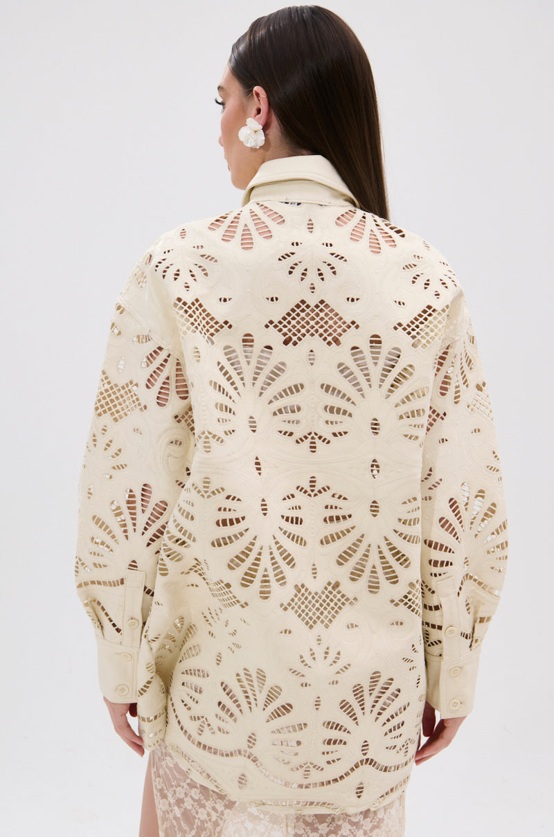 A woman with long brown hair is seen from behind, wearing the YOU FEELIN ME NOW CUTOUT PU BUTTON UP SHIRT in cream, featuring cutout details and floral-inspired lace, paired with large floral earrings against a white backdrop.