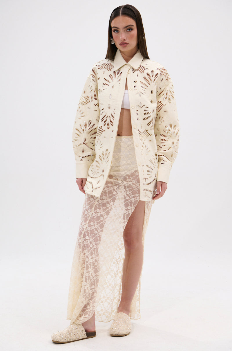 A woman with straight hair and a neutral expression stands against a white background wearing the YOU FEELIN ME NOW CUTOUT PU BUTTON UP SHIRT over a white bralette, paired with a sheer lace maxi skirt and cream slip-on shoes.