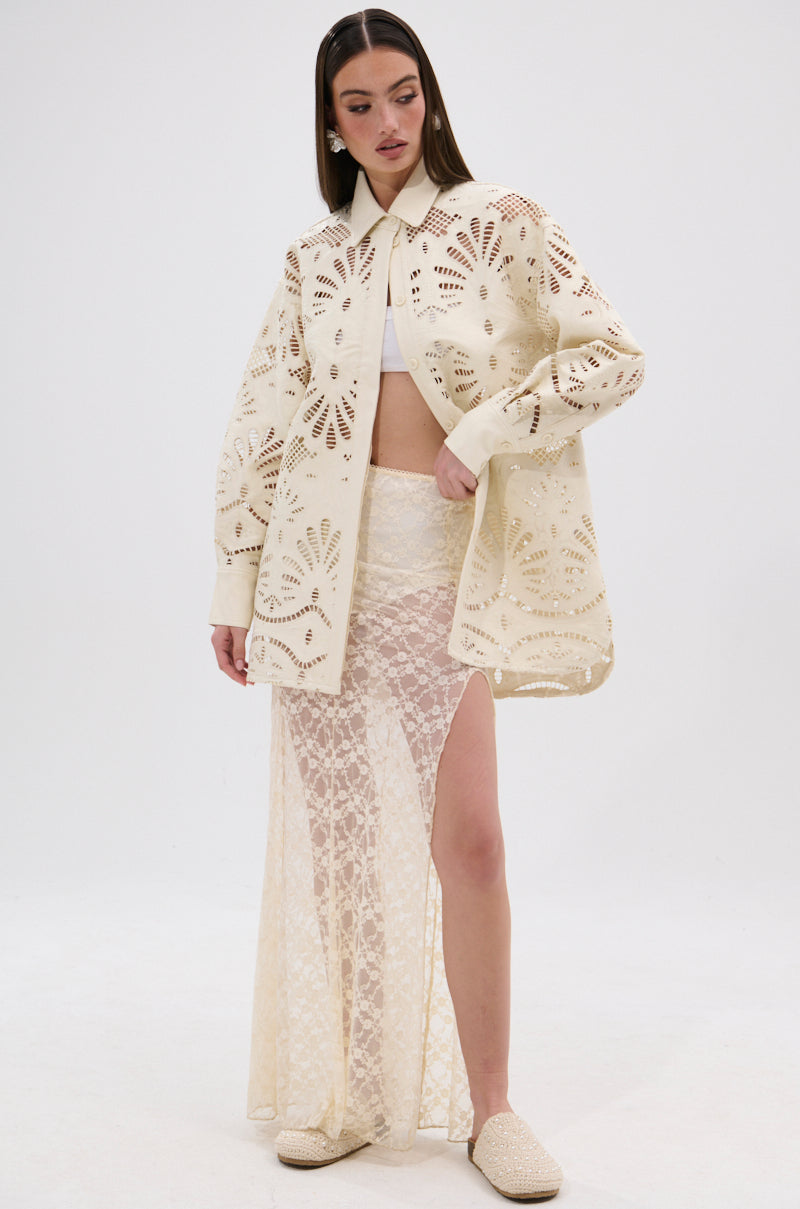 A woman poses in the YOU FEELIN ME NOW CUTOUT PU BUTTON UP SHIRT, styled with a white crop top, sheer lace skirt with high slit, and matching slip-on shoes.