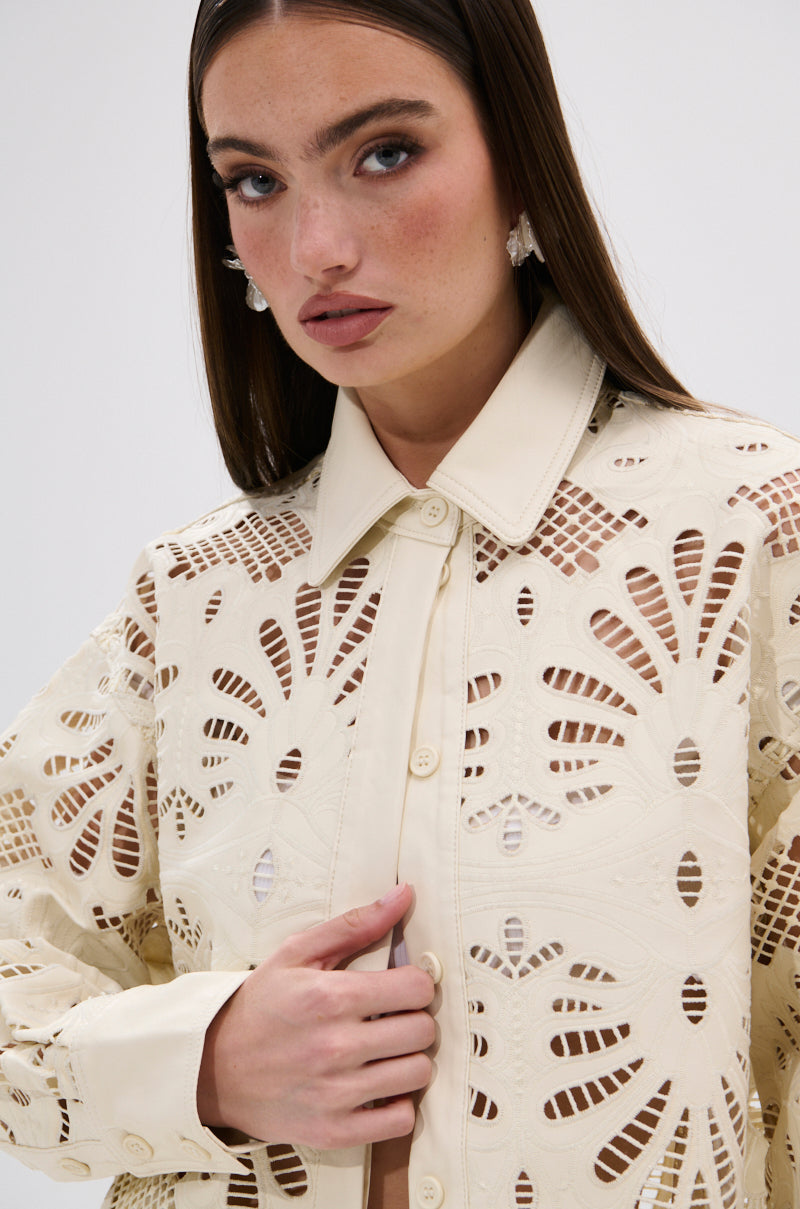 A woman with long brown hair wears the YOU FEELIN ME NOW CUTOUT PU BUTTON UP SHIRT in cream, featuring cutout detailing and floral-inspired patterns. She gazes slightly to the side with a neutral expression.
