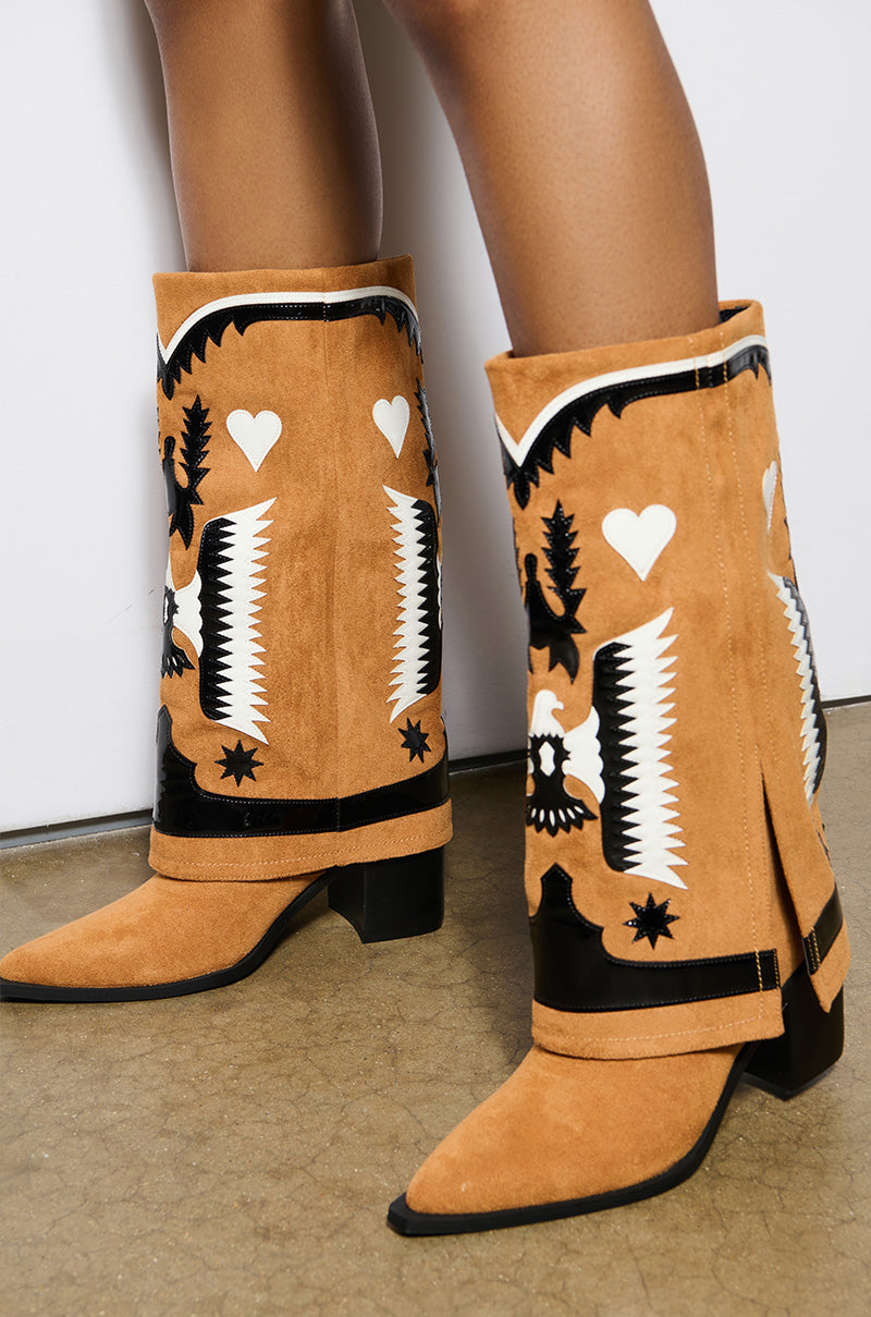 A person wearing the AZALEA WANG HIKARU Brown Faux Suede Western Boot, which boasts black and white decorative patterns with hearts and stars, stands on a concrete floor. These Western boots feature pointed toes and chunky block heels, adding a bold touch to the ensemble.