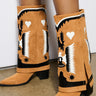 A person wearing the AZALEA WANG HIKARU Brown Faux Suede Western Boot, which boasts black and white decorative patterns with hearts and stars, stands on a concrete floor. These Western boots feature pointed toes and chunky block heels, adding a bold touch to the ensemble.