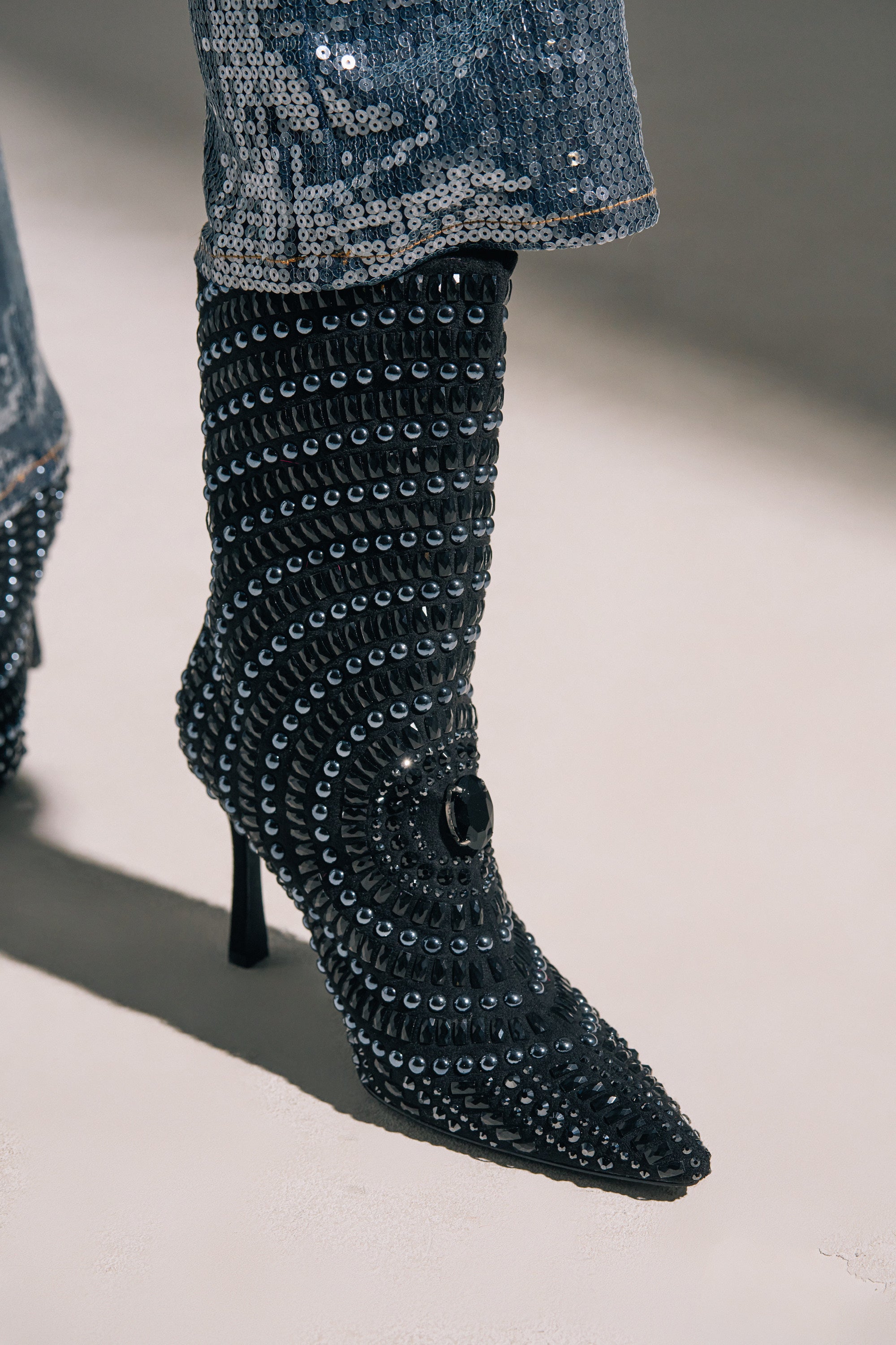 A close-up of the AZALEA WANG HILLEL PEWTER RHINESTONE BOOTIE, a high-heeled, pointed-toe stiletto covered in black and silver rhinestones forming a circular pattern, paired with sequined pants.