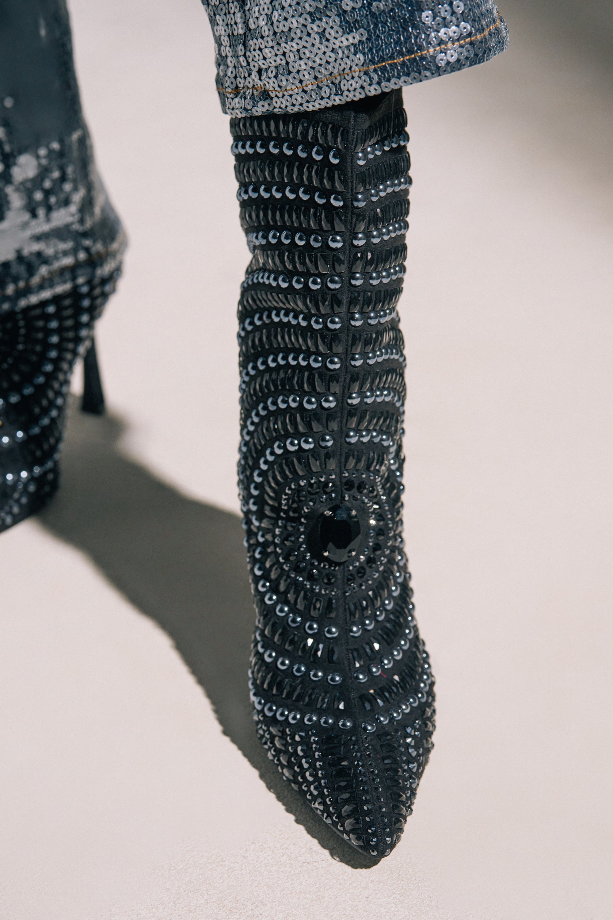 A close-up of the AZALEA WANG HILLEL PEWTER RHINESTONE BOOTIE, featuring a stiletto heel and dazzling black studs and sequins, paired with sequined pants. The faux suede texture and intricate detailing make this bootie a bold statement piece.