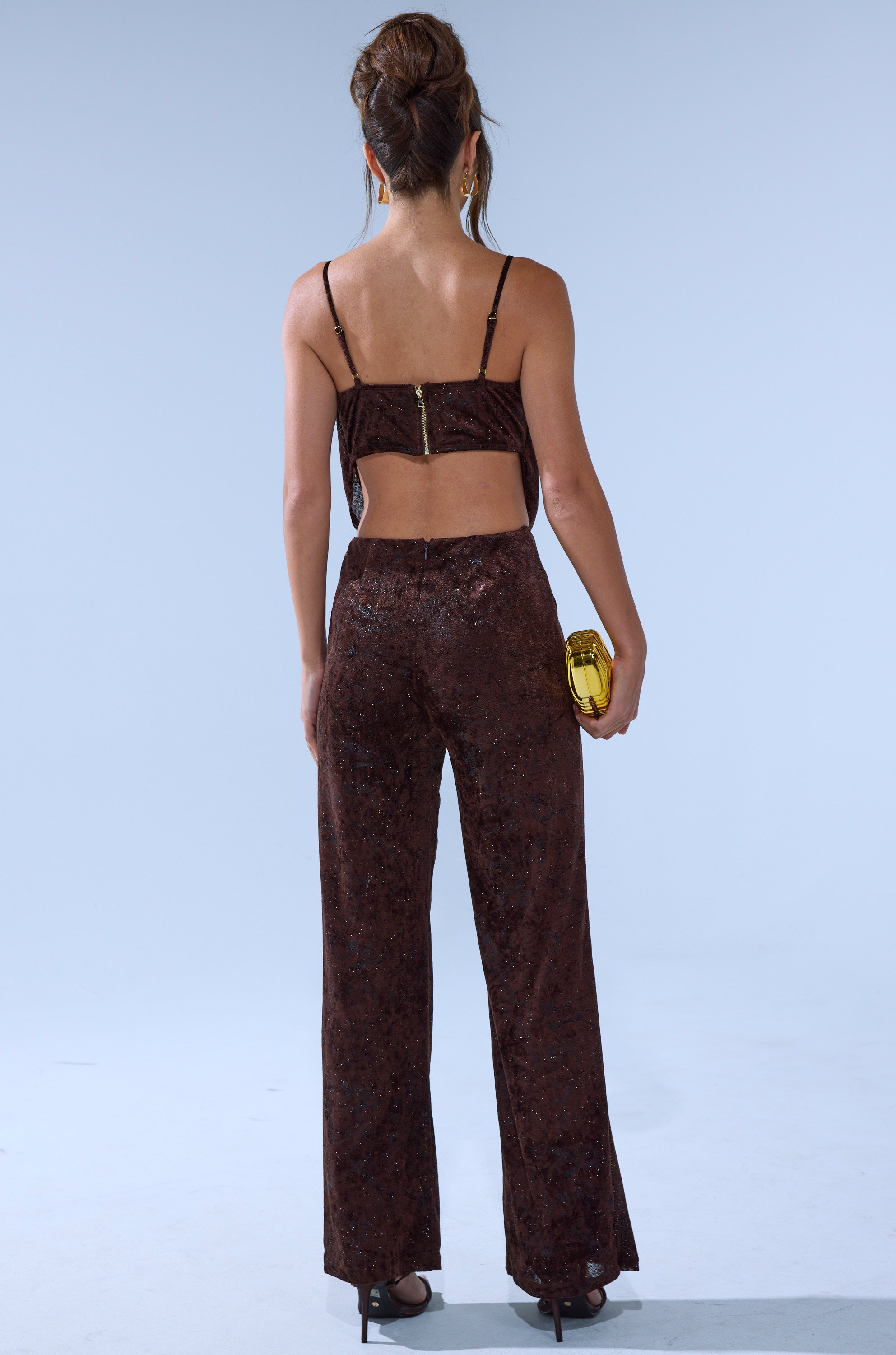 A woman faces away, wearing the MELANY COWL NECK JUMPSUIT in dark brown velvet with high-waisted pants. She holds a small yellow clutch, wears high heels and earrings, has her hair up, and stands against a plain light blue background.