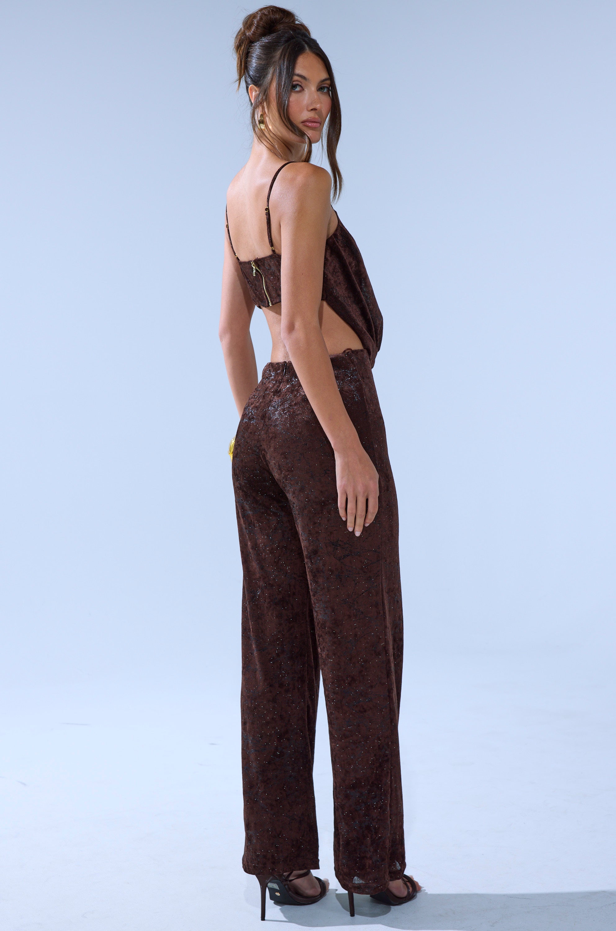 A woman stands sideways, looking over her shoulder, wearing the MELANY COWL NECK JUMPSUIT in dark brown velvet with wide legs and high heels against a plain light background.