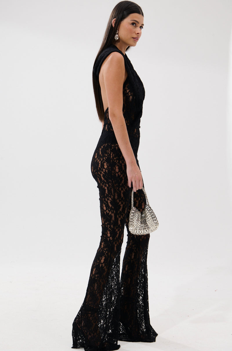 A woman poses sideways in the JUMPIN AROUND LACE JUMPSUIT, featuring a sleeveless design, black lace, flared legs, and a plunging neckline. She accessorizes with a silver handbag and drop earrings; her straight hair contrasts the plain white background.