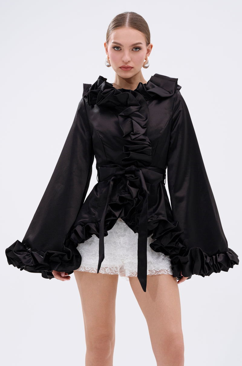 A woman wears the GARDEN PARTY WRAP BLAZER—an eye-catching black blazer with flared sleeves and a tie waist—over a short white lace skirt, standing against a plain white background. 0