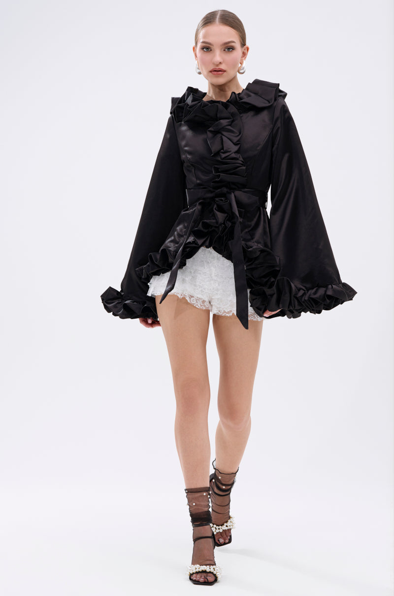 A woman stands against a plain white background, wearing the chic GARDEN PARTY WRAP BLAZER over a black ruffled long-sleeve top with a bow, white lace shorts, and strappy pearl-detailed high-heeled sandals. 9