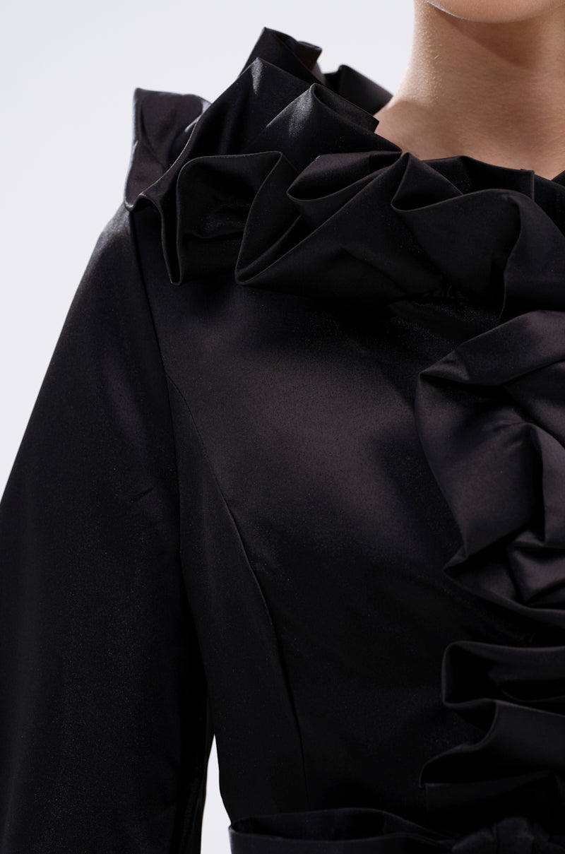 Close-up of a person wearing the GARDEN PARTY WRAP BLAZER, featuring black satin fabric and dramatic ruffled detailing at the neckline and chest, with only the lower face and upper body partially visible as the blazer catches the light. 10