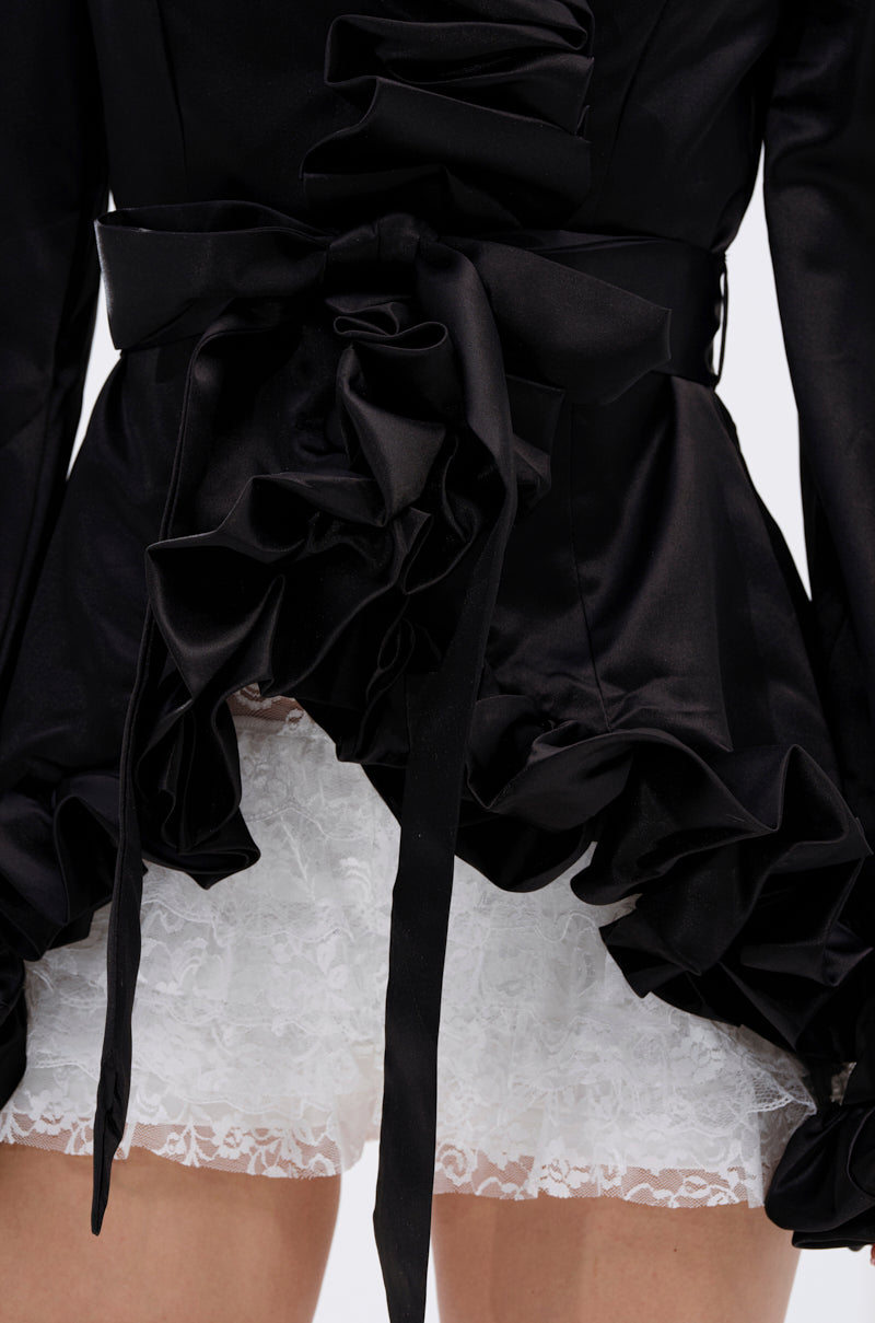 Close-up of a person in a black ruffled dress styled with the GARDEN PARTY WRAP BLAZER and tied belt, revealing white lace-trimmed shorts. The image highlights the contrasting fabrics and textures of the outfit. 11