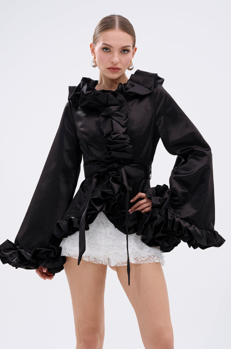 A woman models the GARDEN PARTY WRAP BLAZER, a black satin piece with ruffled trim and wide bell sleeves, paired with white lace shorts. She wears statement earrings and has slicked-back hair, posing confidently against a plain backdrop. 1