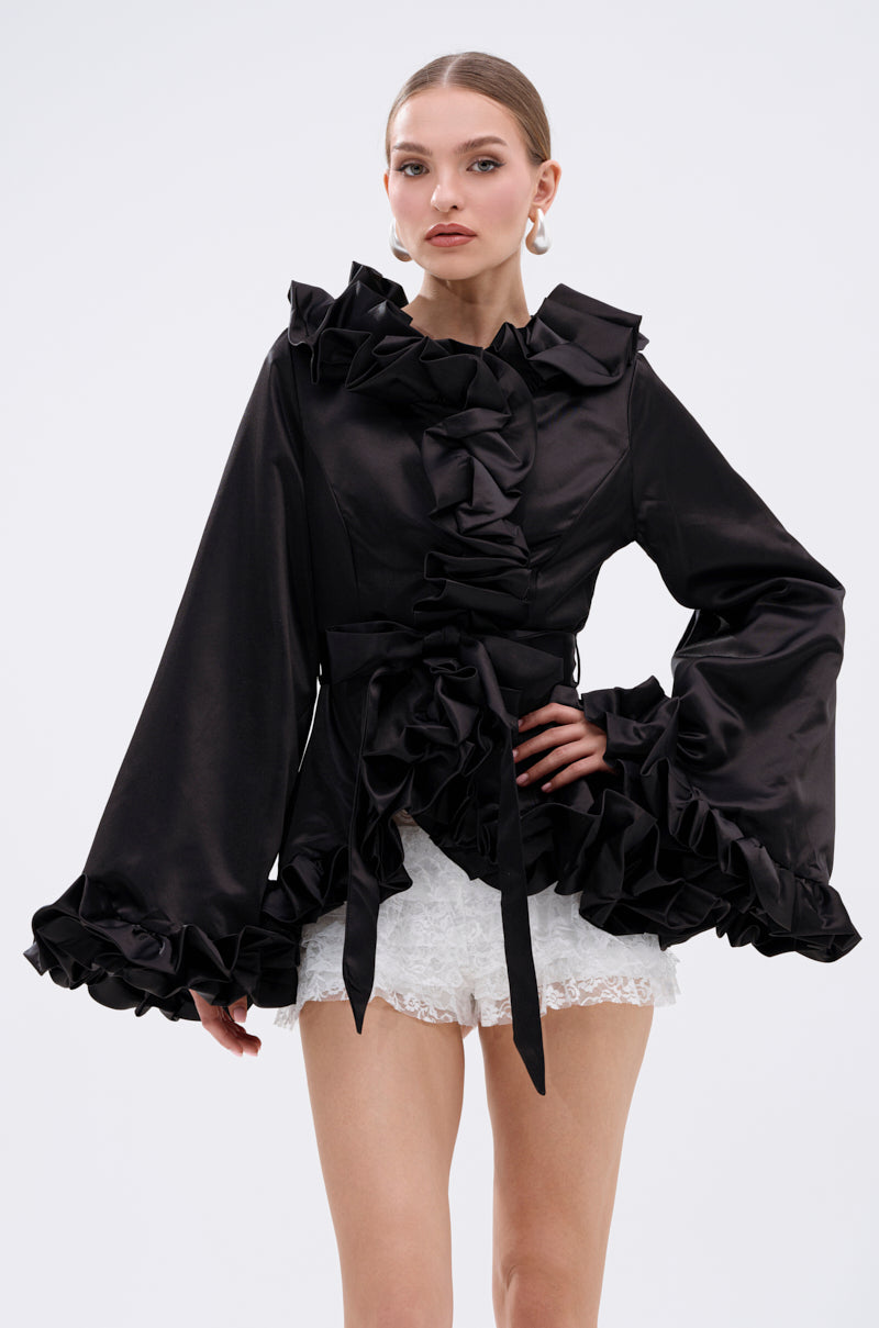 A woman poses confidently in the GARDEN PARTY WRAP BLAZER—black, long-sleeved with ruffles and a tie belt—paired with white textured shorts against a plain light background. 2