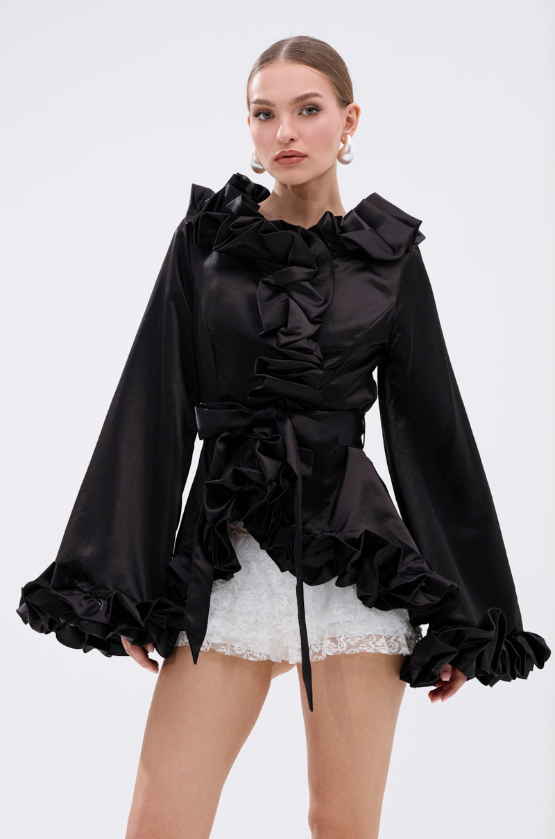 A woman stands against a plain light background wearing the GARDEN PARTY WRAP BLAZER, a black, long-sleeved ruffle blazer with a tie belt, paired with white lace shorts. 3