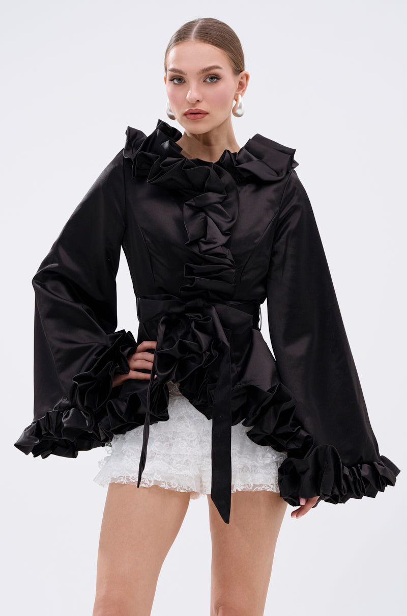 A woman stands against a plain background wearing the GARDEN PARTY WRAP BLAZER, a black long-sleeve blazer with ruffled edges and a tie belt, paired with white lace shorts. 4