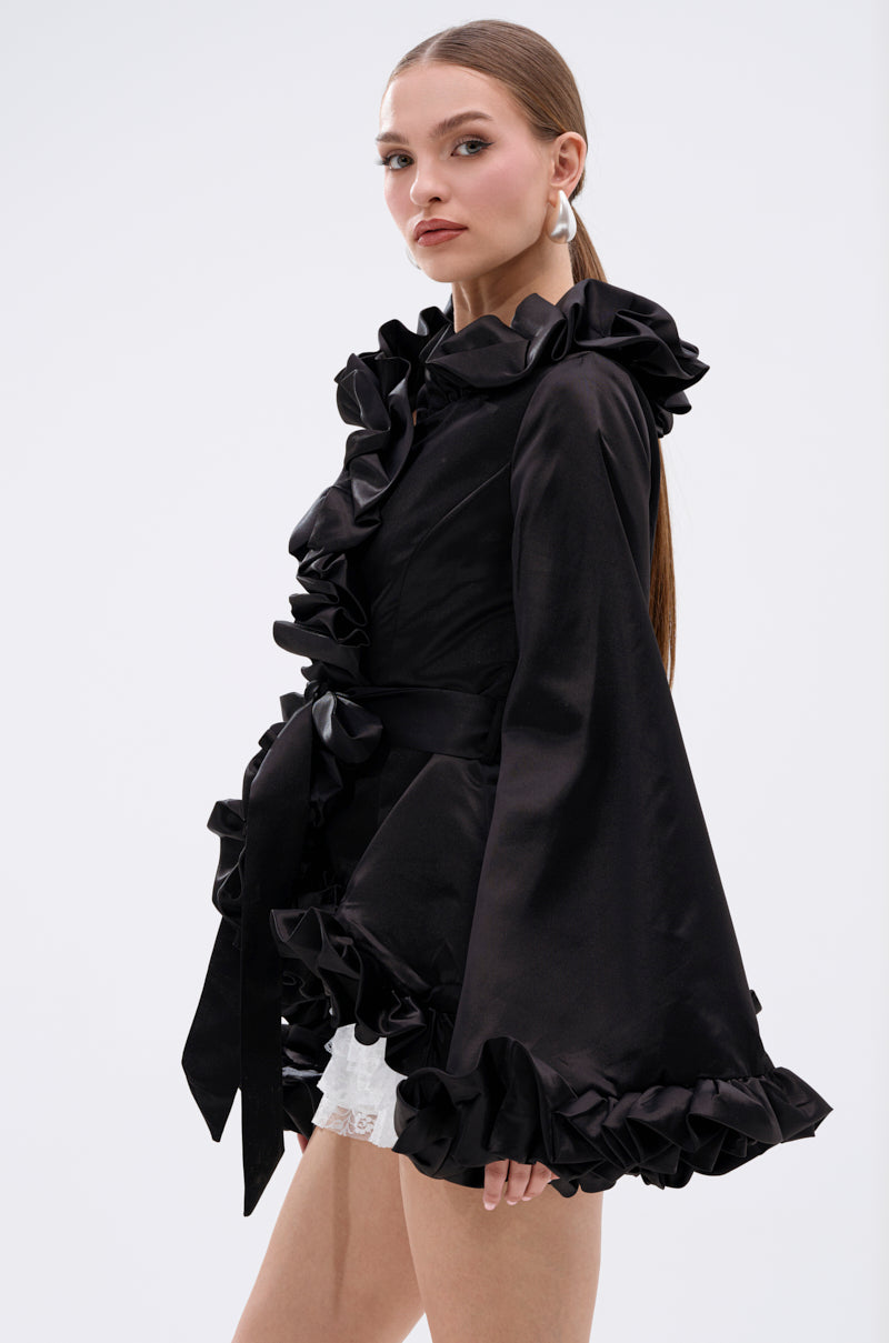 A woman with long straight hair confidently models the GARDEN PARTY WRAP BLAZER, featuring dramatic ruffled trim on the collar, sleeves, and hem, paired with white lace shorts against a plain white background. 6
