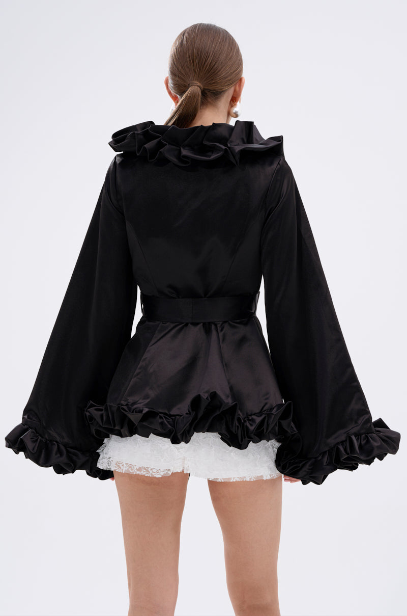 A woman with a low ponytail faces away, wearing the GARDEN PARTY WRAP BLAZER—black with dramatic ruffled collar and sleeves—over a short white lace skirt. The background is plain and light-colored. 7