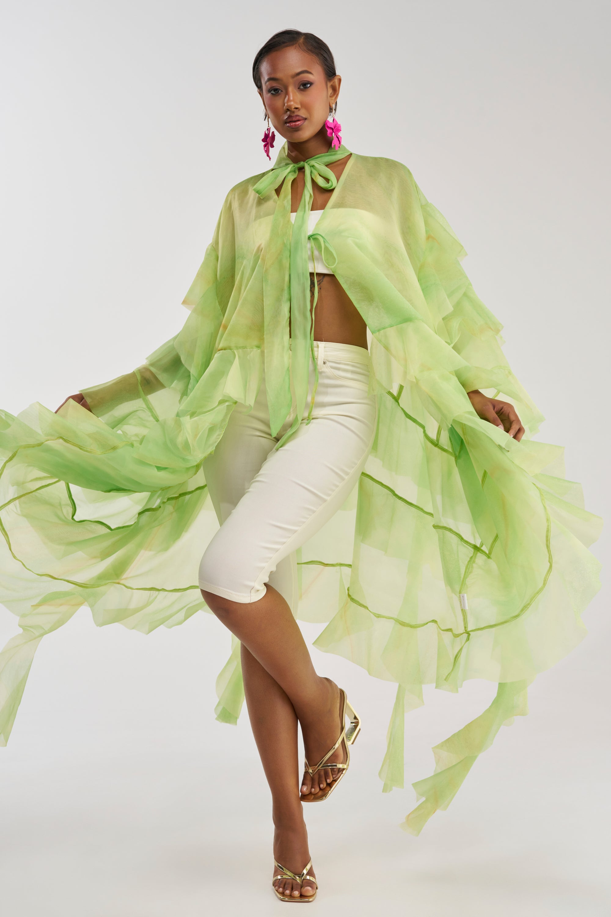 A woman poses confidently against a plain, light background, wearing the IN BLOOM FLORAL TOP IN GREEN MULTI—a sheer, flowing chiffon blouse with ruffles—paired with white cropped pants, gold heels, and bright pink earrings.